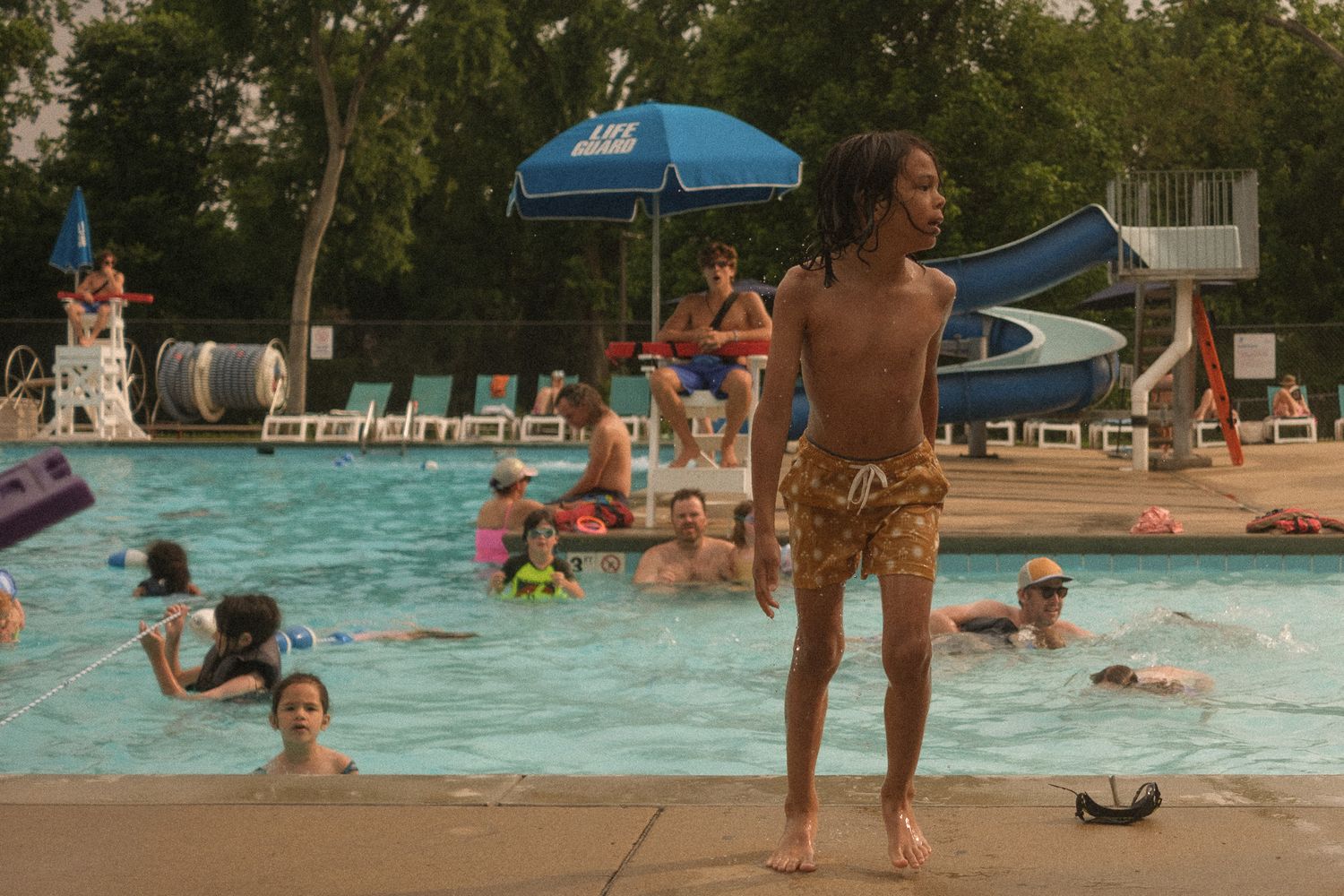 Young boy standing poolside at a busy outdoor community swimming pool in summer.