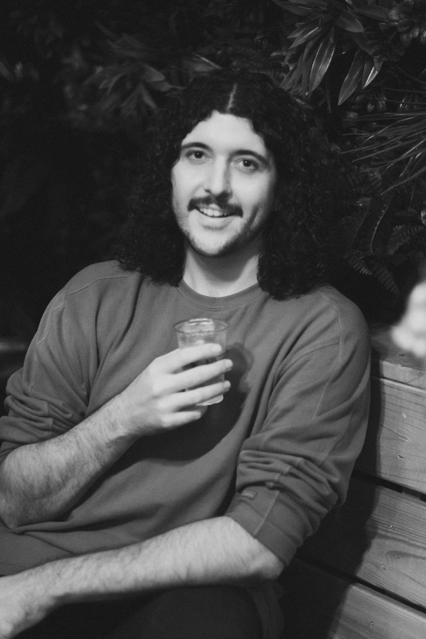 Black and white portrait of a young man with long curly hair and mustache holding a drink while sitting outdoors.