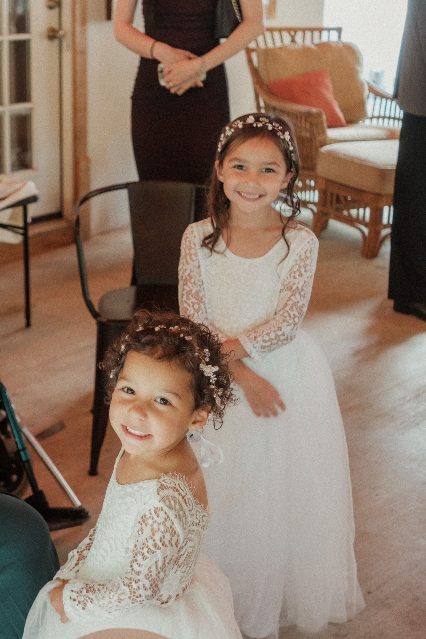 Two young flower girls in white lace dresses smile joyfully during wedding preparations indoors.