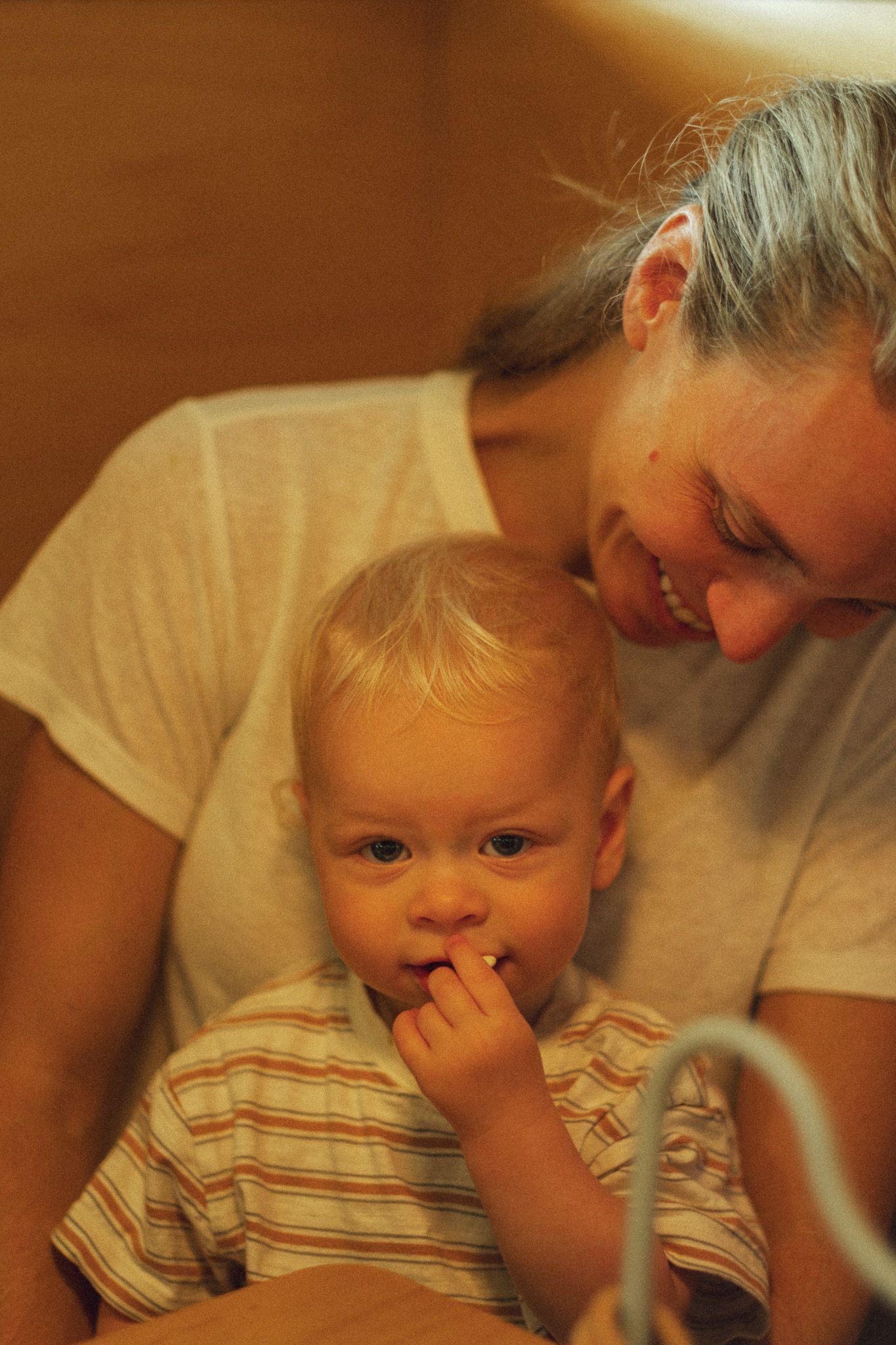 Smiling mother holding her blonde toddler who is putting fingers in mouth.