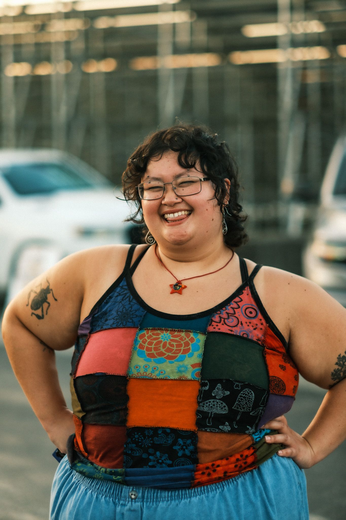 Plus-size woman with short dark hair smiling broadly, wearing a colorful patchwork tank top with hands on hips.