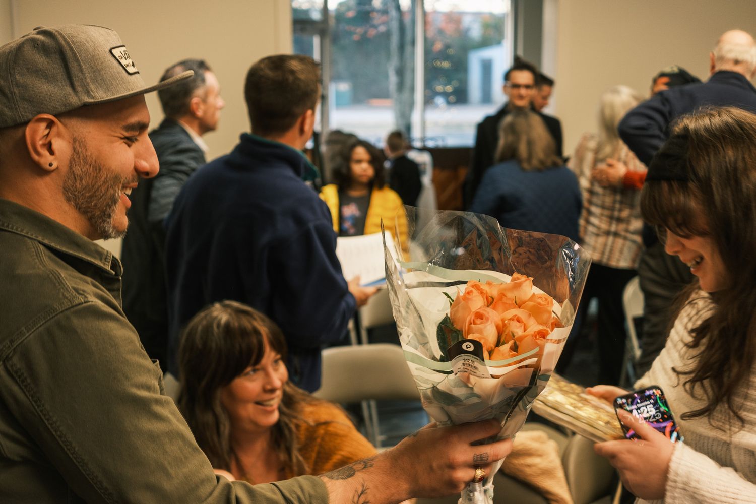 A man in a cap presents a bouquet of orange flowers to a smiling woman at an indoor gathering.