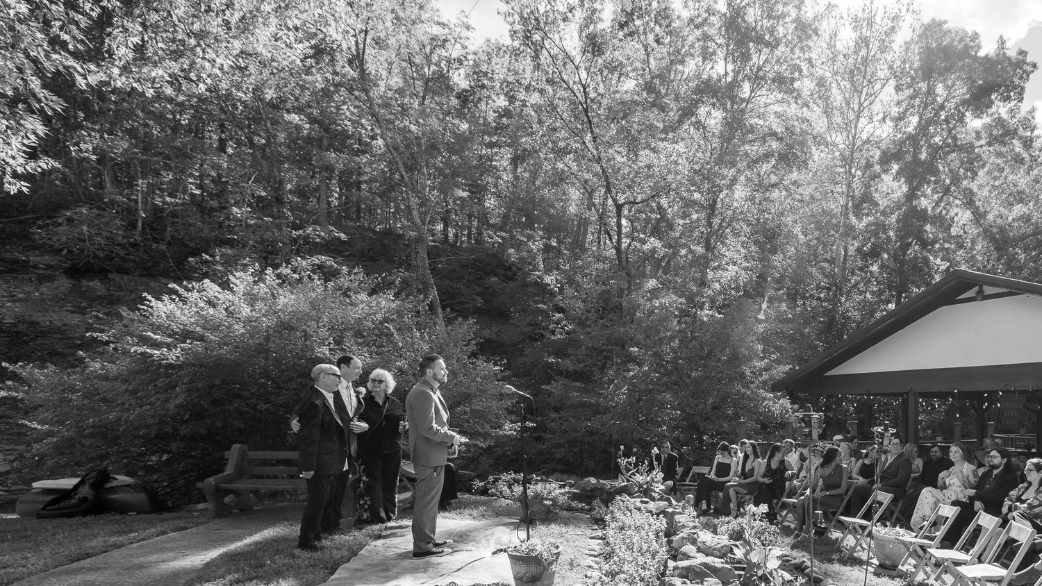 Black and white photo of outdoor wedding ceremony with guests seated among lush trees and natural landscape.
