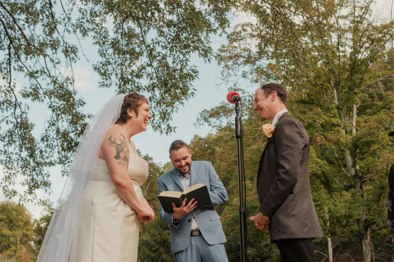 Outdoor wedding ceremony with bride, groom, and officiant reading vows beneath green trees.