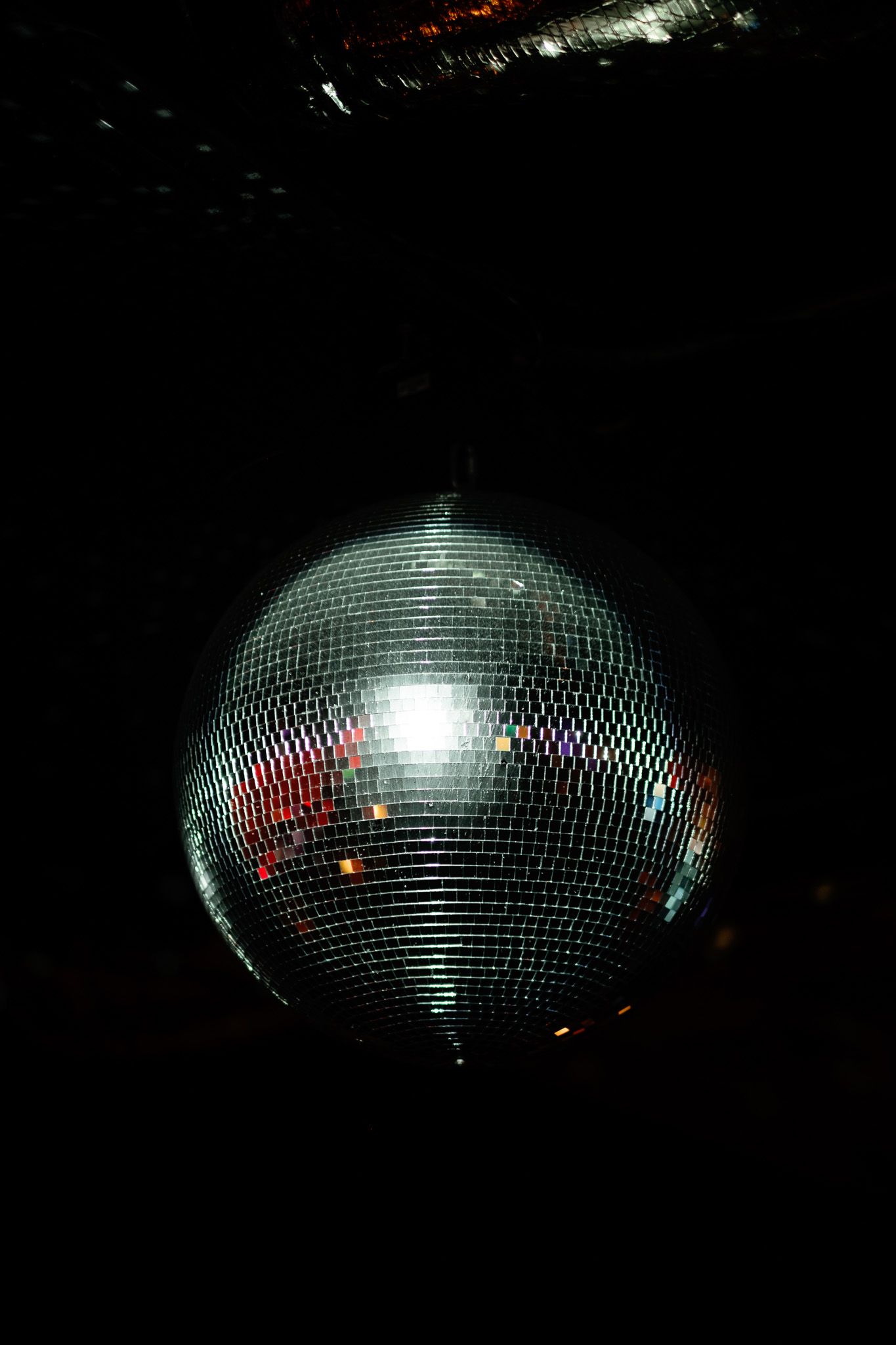 Shimmering silver disco mirror ball hanging from a dark ceiling reflecting colorful light.