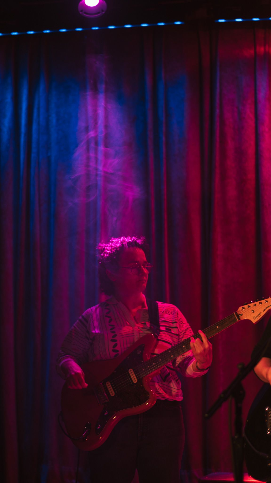 Guitarist performing on stage under purple and red lights with smoke and blue curtain backdrop.