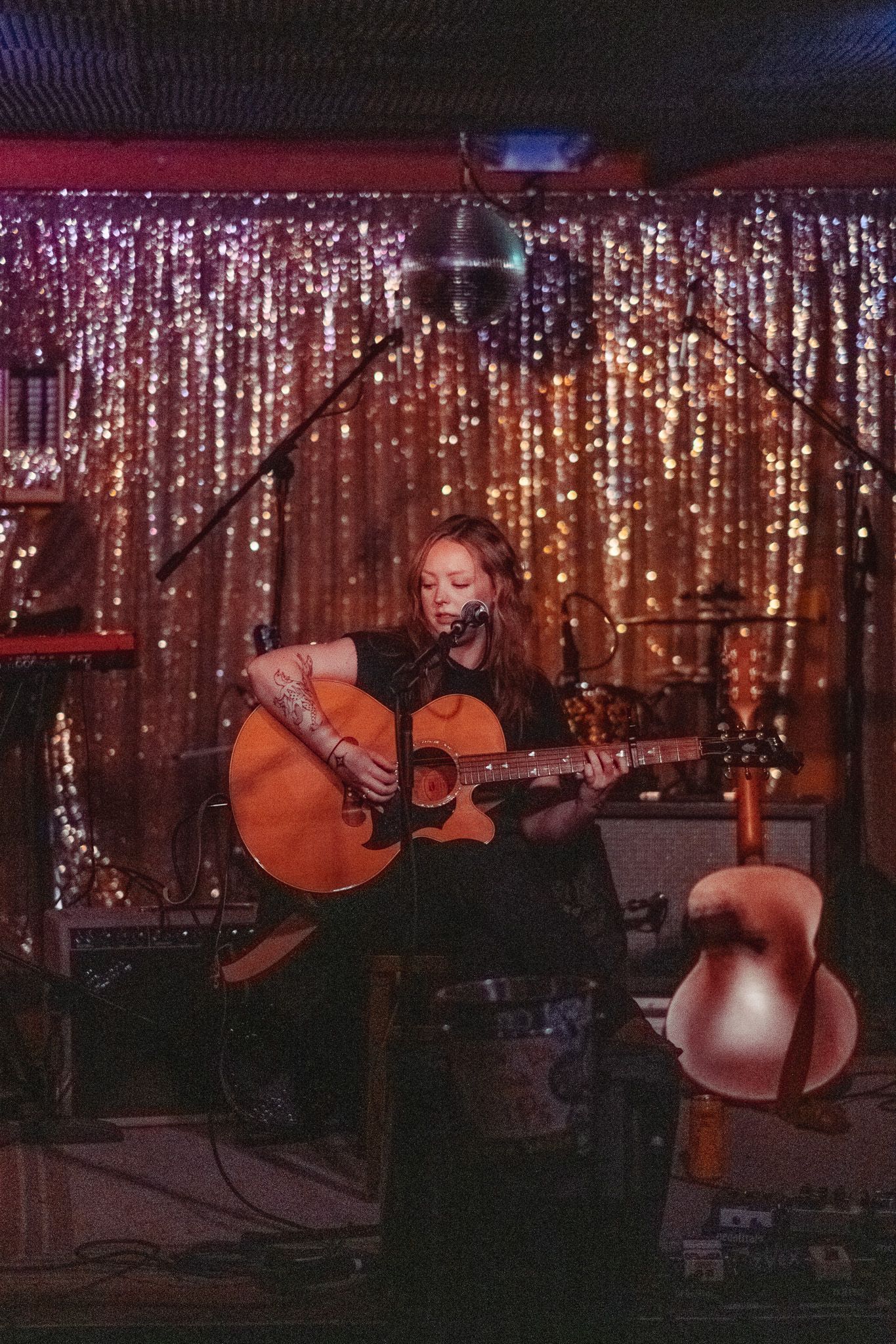 Female singer-songwriter performing on stage with acoustic guitar before a sparkling gold curtain backdrop.