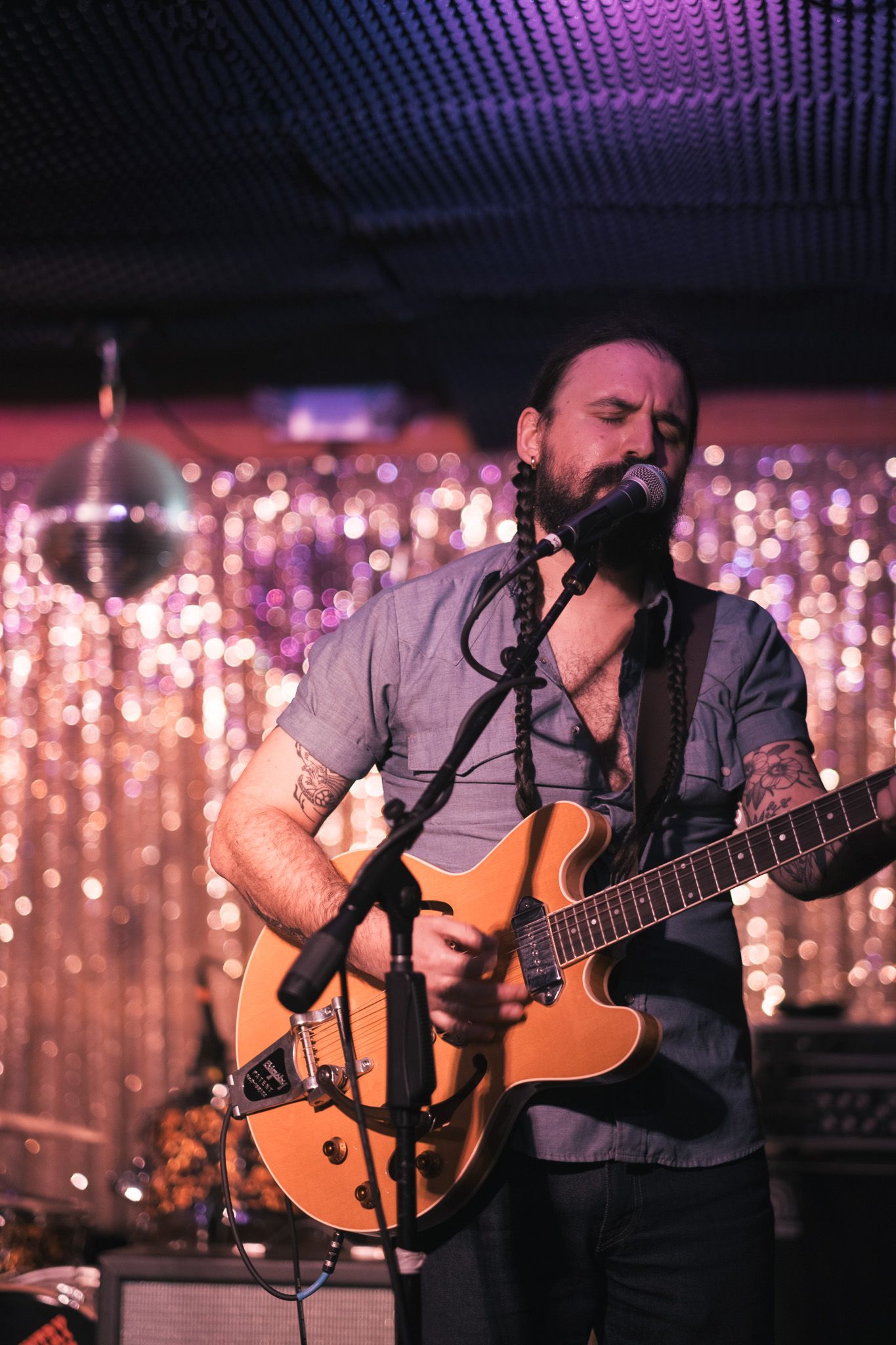 Bearded guitarist with tattoos performs passionately at a microphone on a glittery stage.