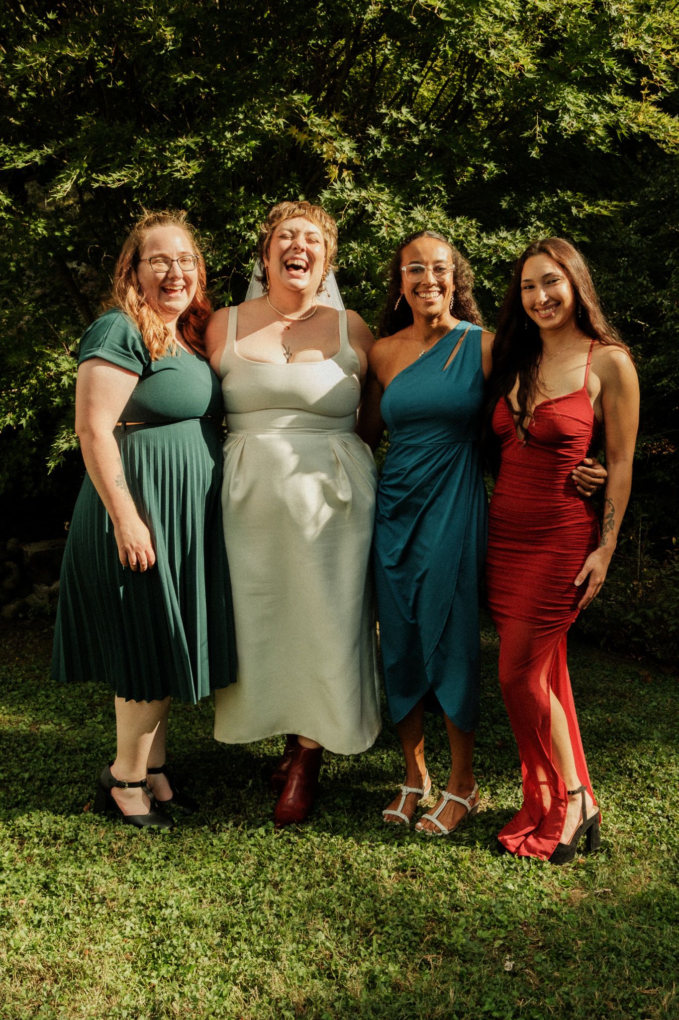 Bride in white satin dress laughing with three bridesmaids in colorful gowns outdoors.
