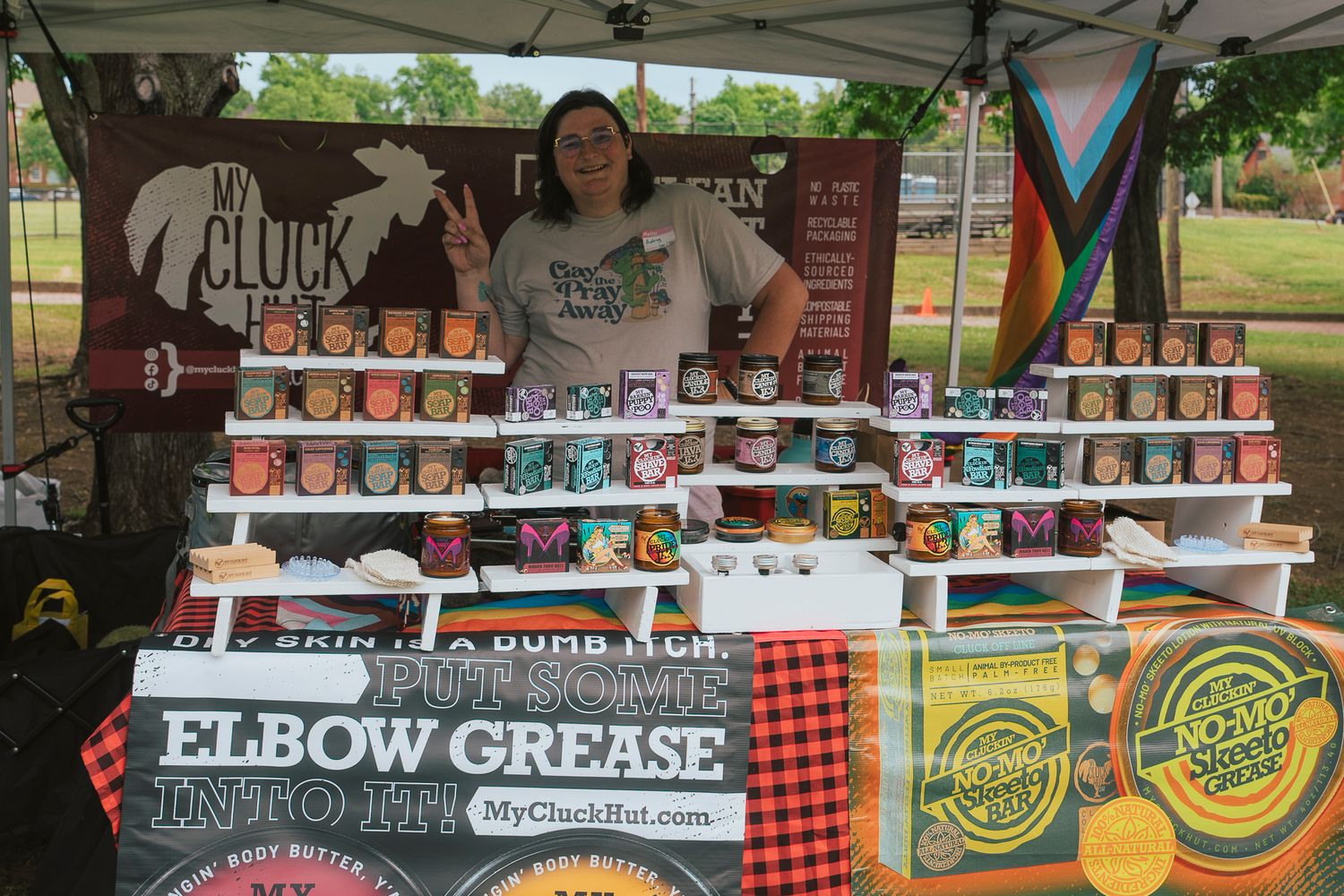 Woman vendor standing behind colorful candle display at outdoor market booth with My Cluck Hut branding.