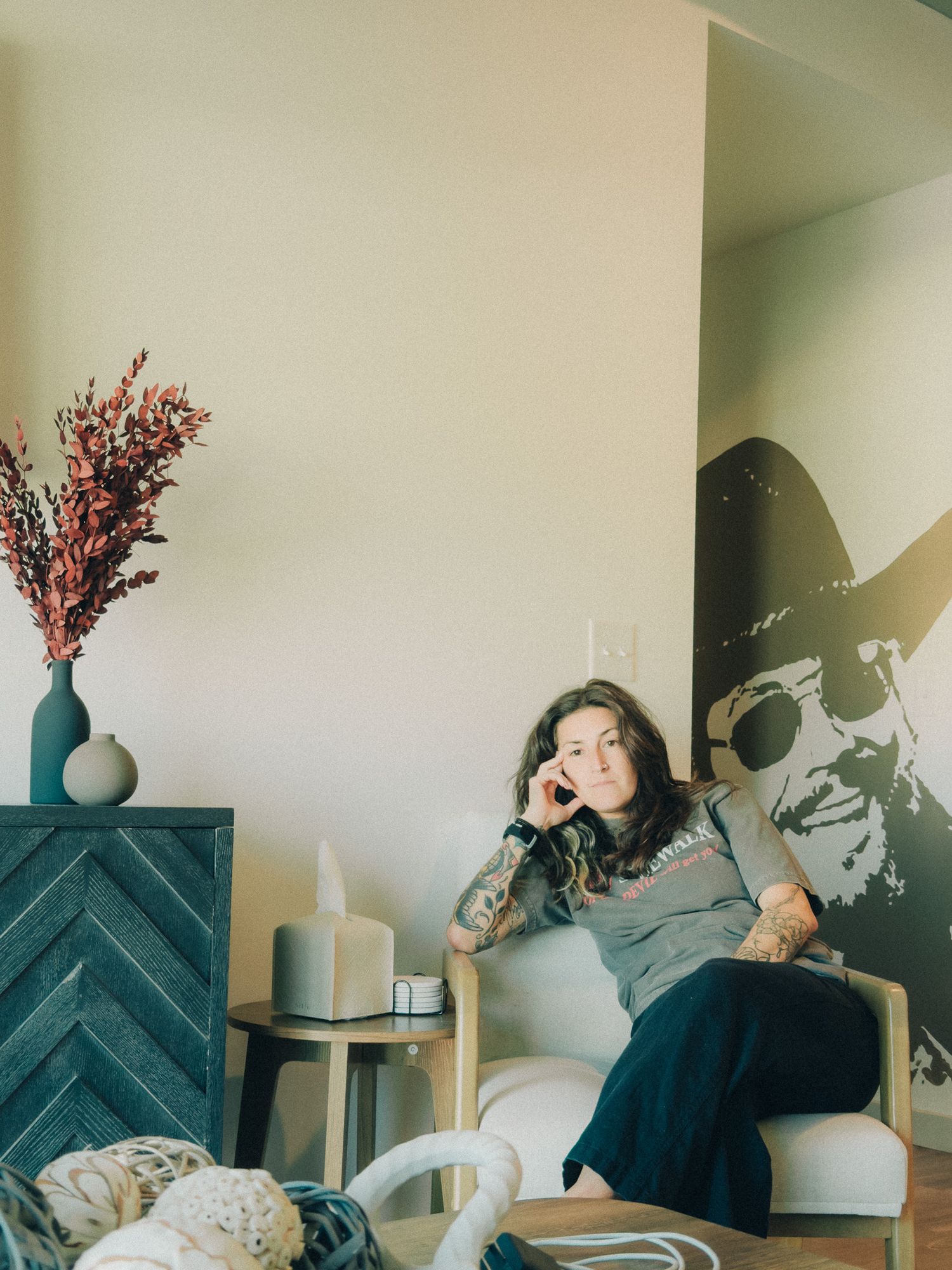 Young woman relaxing in a chair beside a vase of dried flowers with a large wall mural behind her.