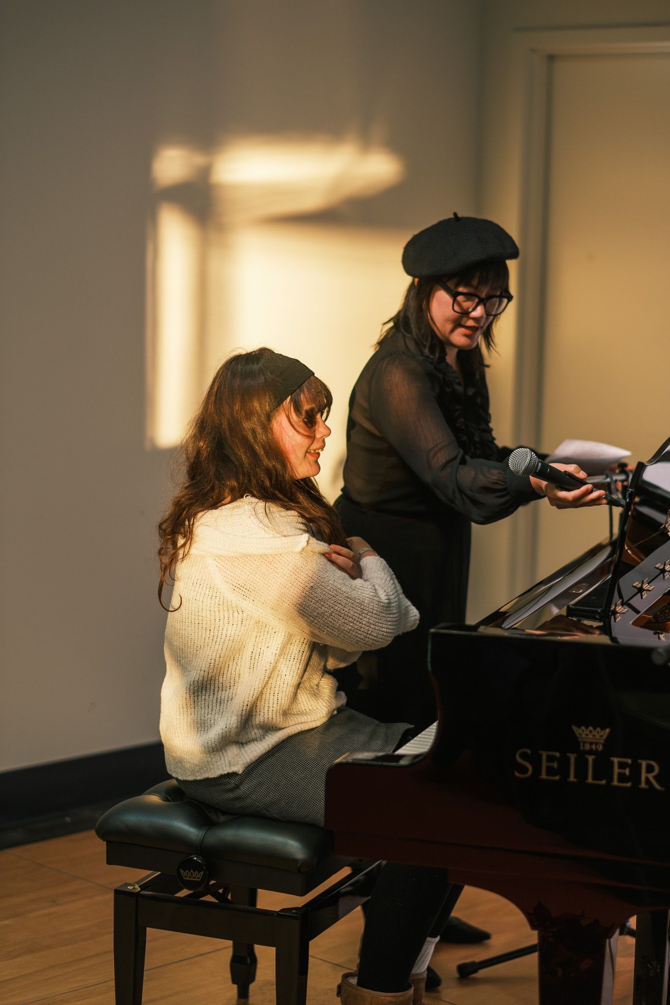 Two women at a Seiler grand piano in warm light, one seated playing while the other stands holding a microphone.