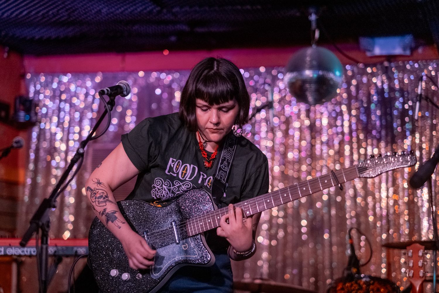 Tattooed female guitarist plays electric guitar on stage with a glittery silver curtain backdrop.