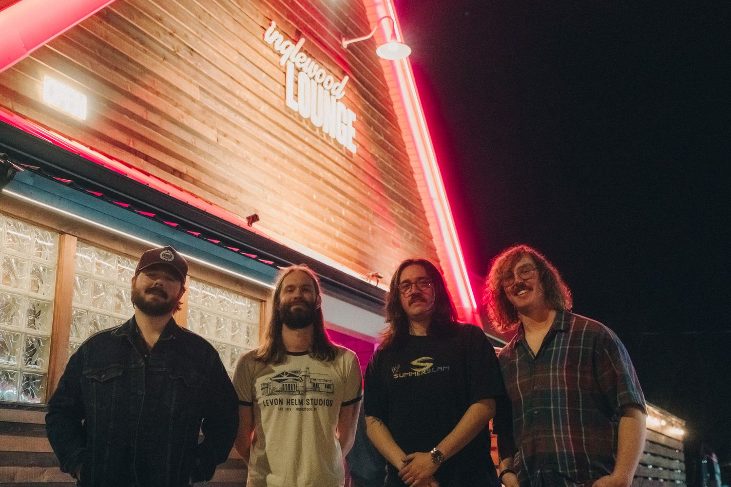 Four-member rock band posing outside Englewood Lounge venue under pink neon lights at night.