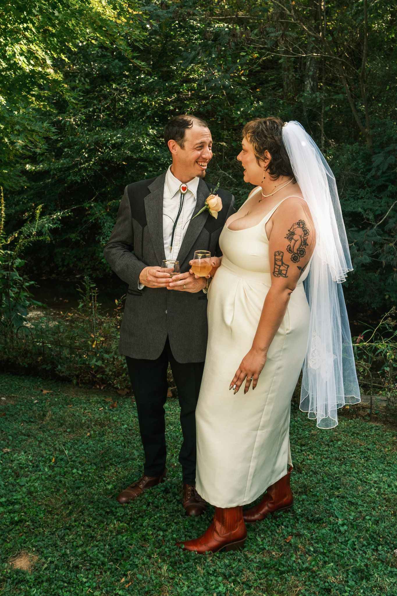 Bride and groom share a joyful moment outdoors, holding drinks and smiling at each other on their wedding day.