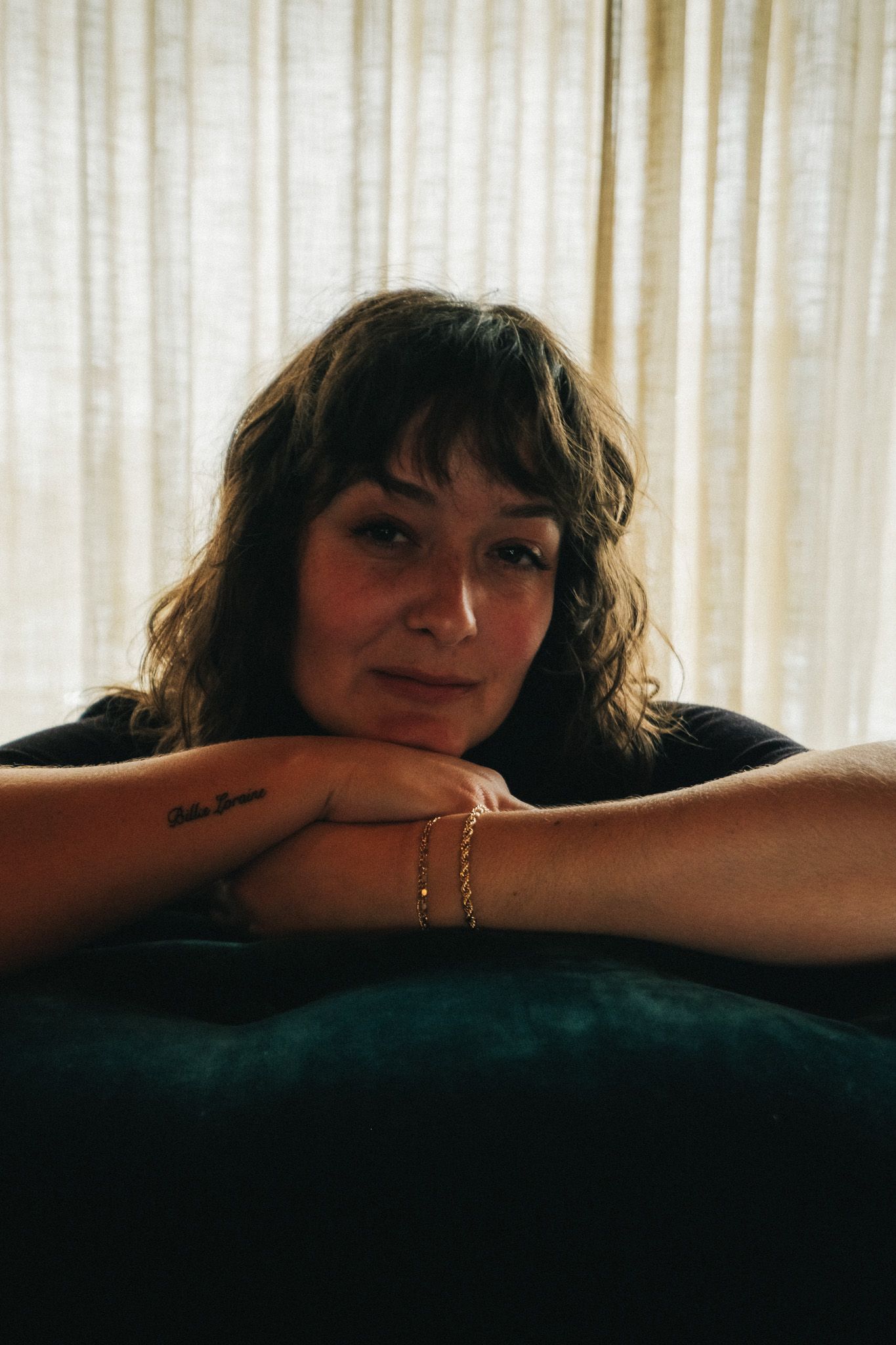 Woman with wavy hair resting her chin on her arms over a teal velvet sofa near curtains.