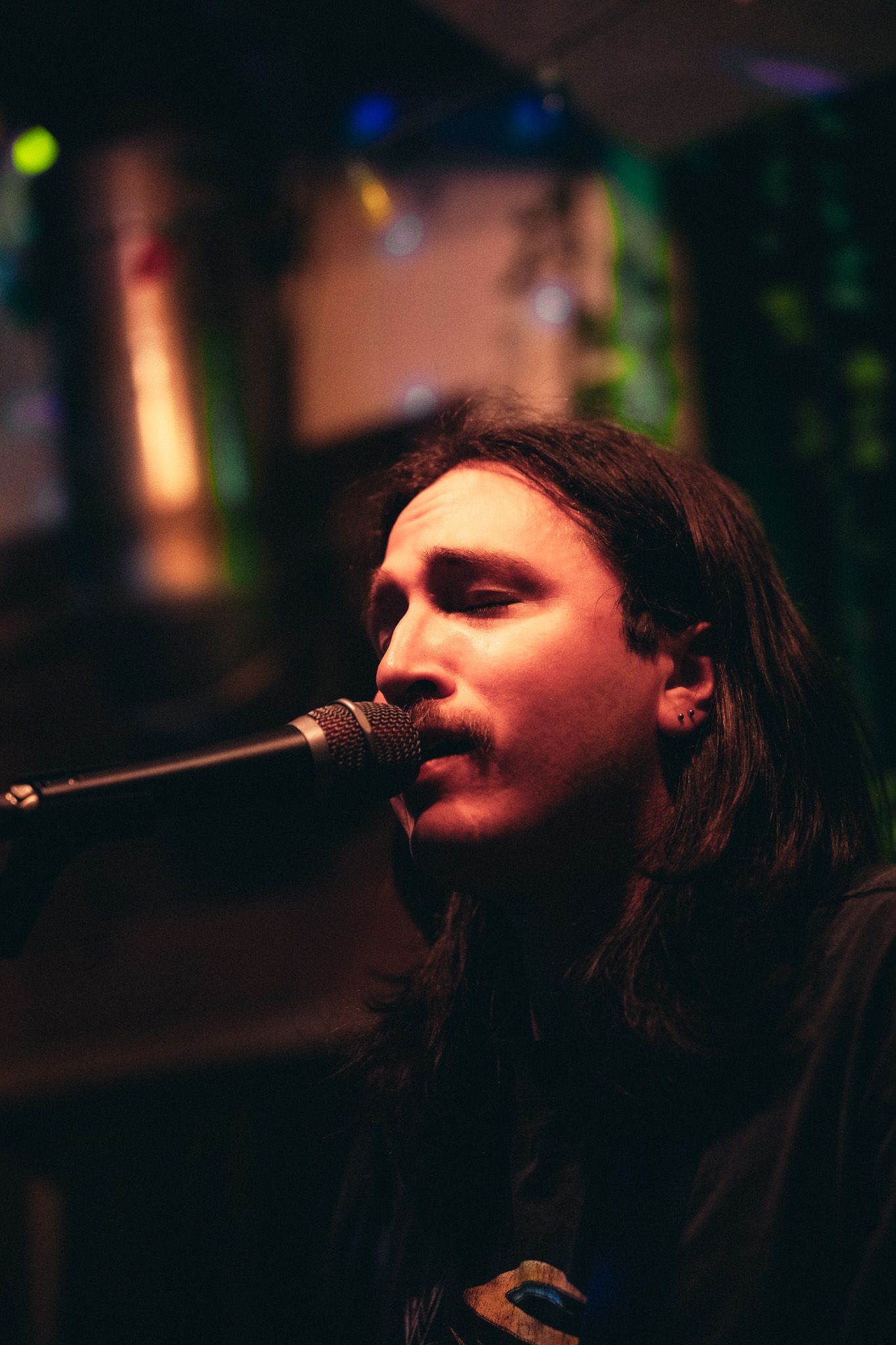 Young male musician with long hair and mustache singing into a microphone under colorful stage lighting.