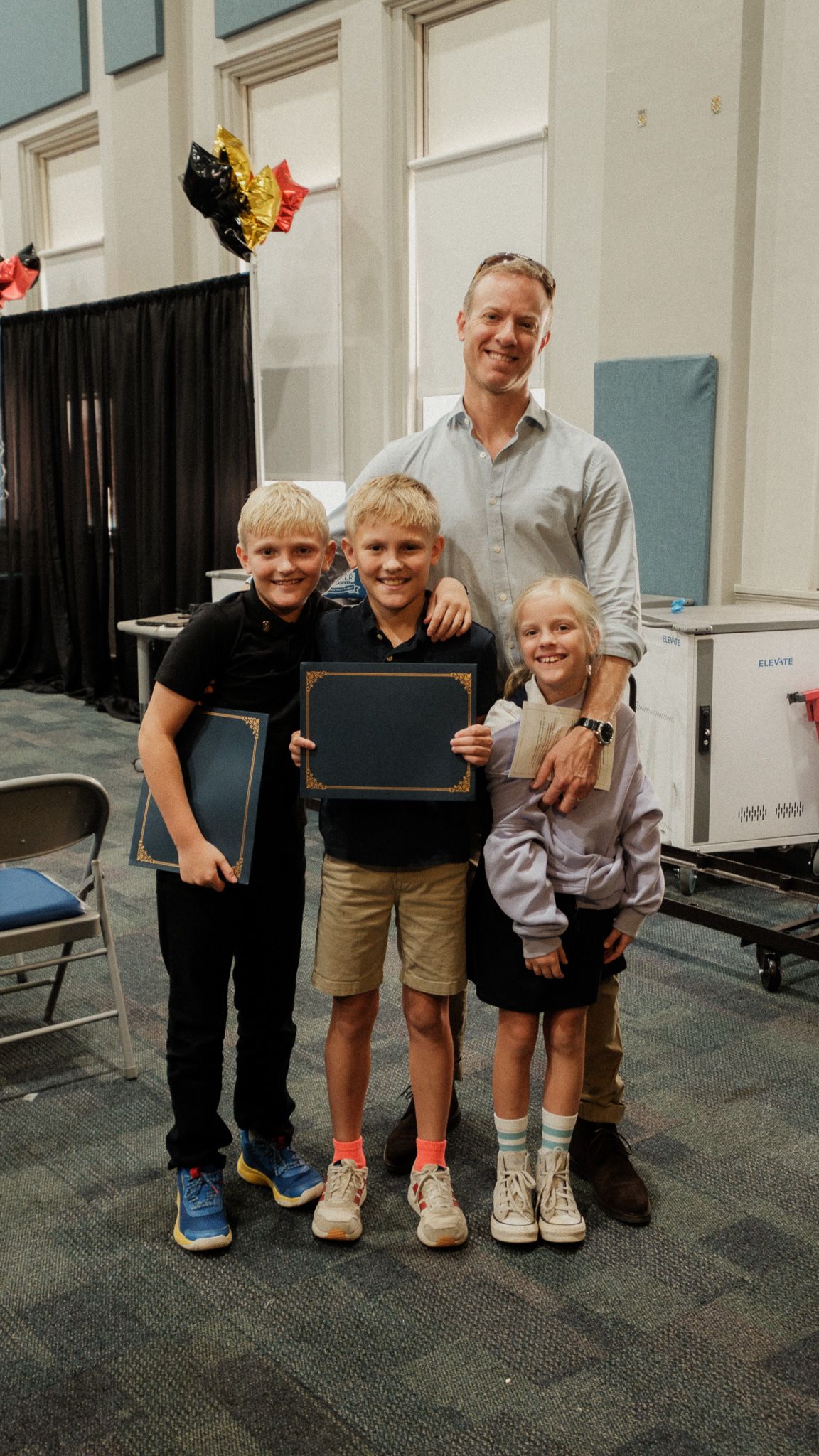 Proud father posing with three children holding award certificates at a school ceremony.