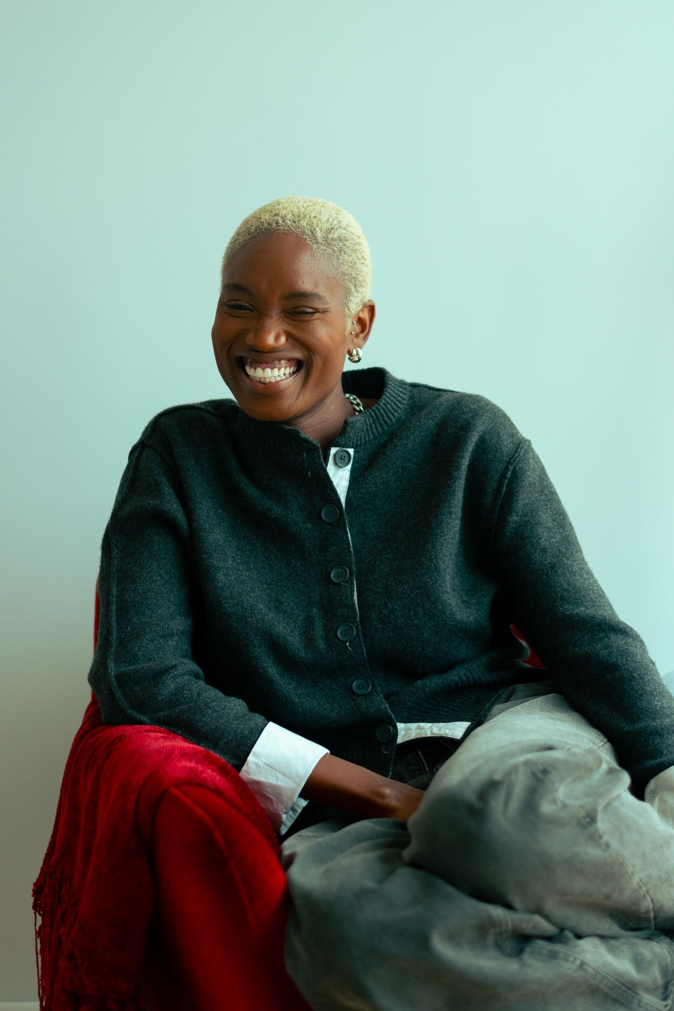 Joyful woman with short blonde hair wearing a dark cardigan sitting against a light blue wall.