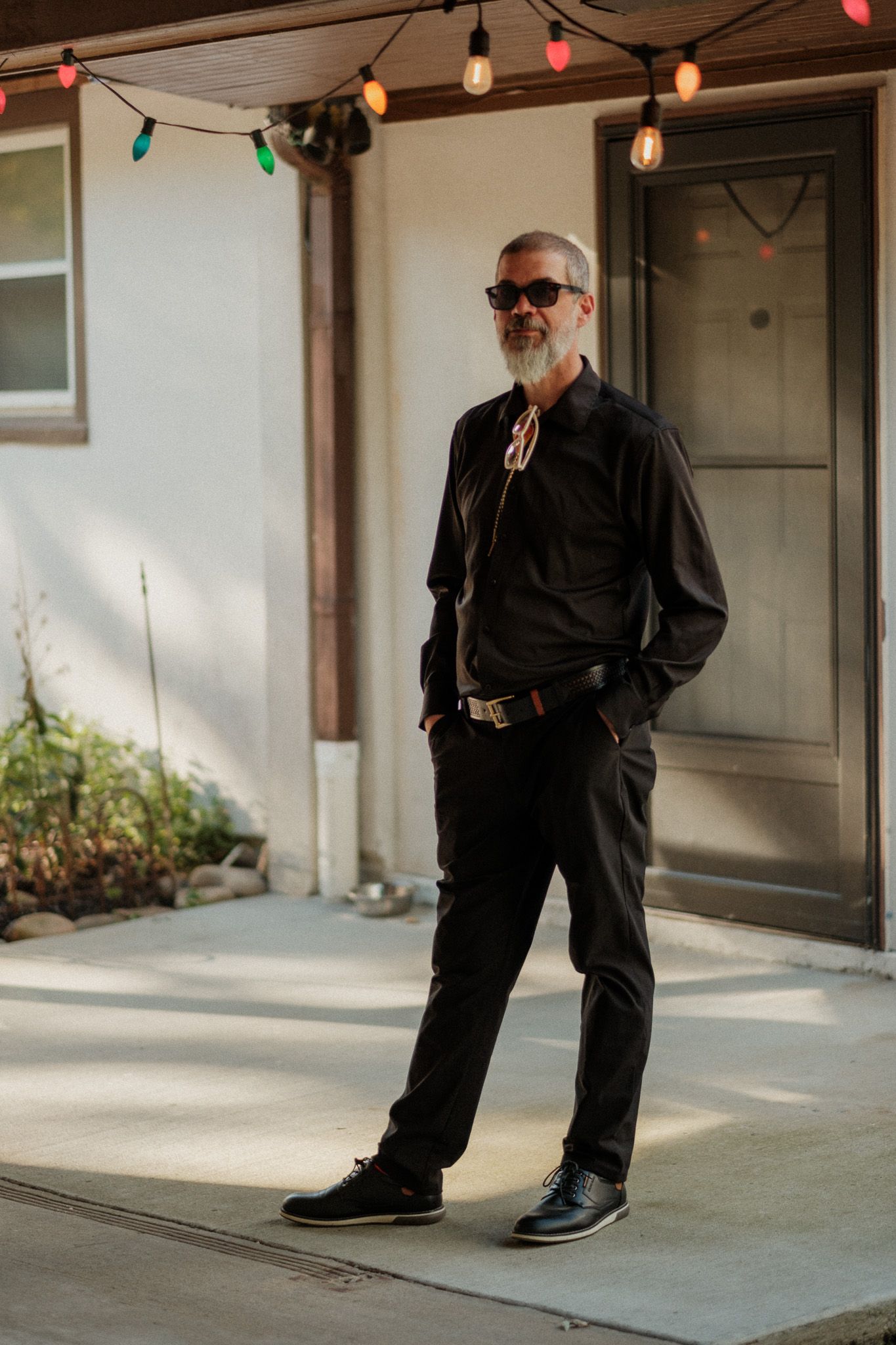 Stylish older man with gray beard wearing all-black outfit and sunglasses standing under string lights.