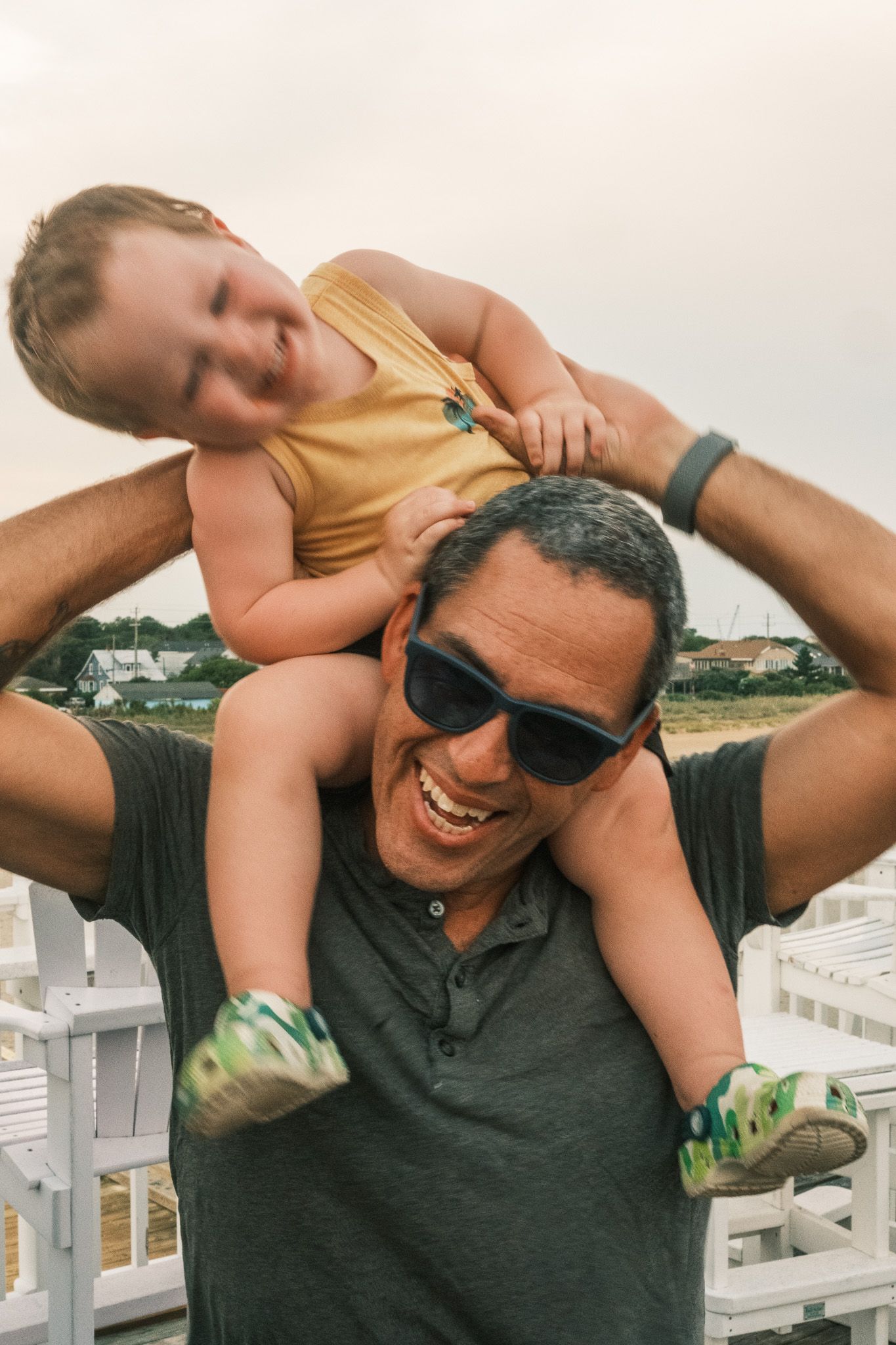 Smiling father carrying laughing toddler on shoulders at a coastal location.