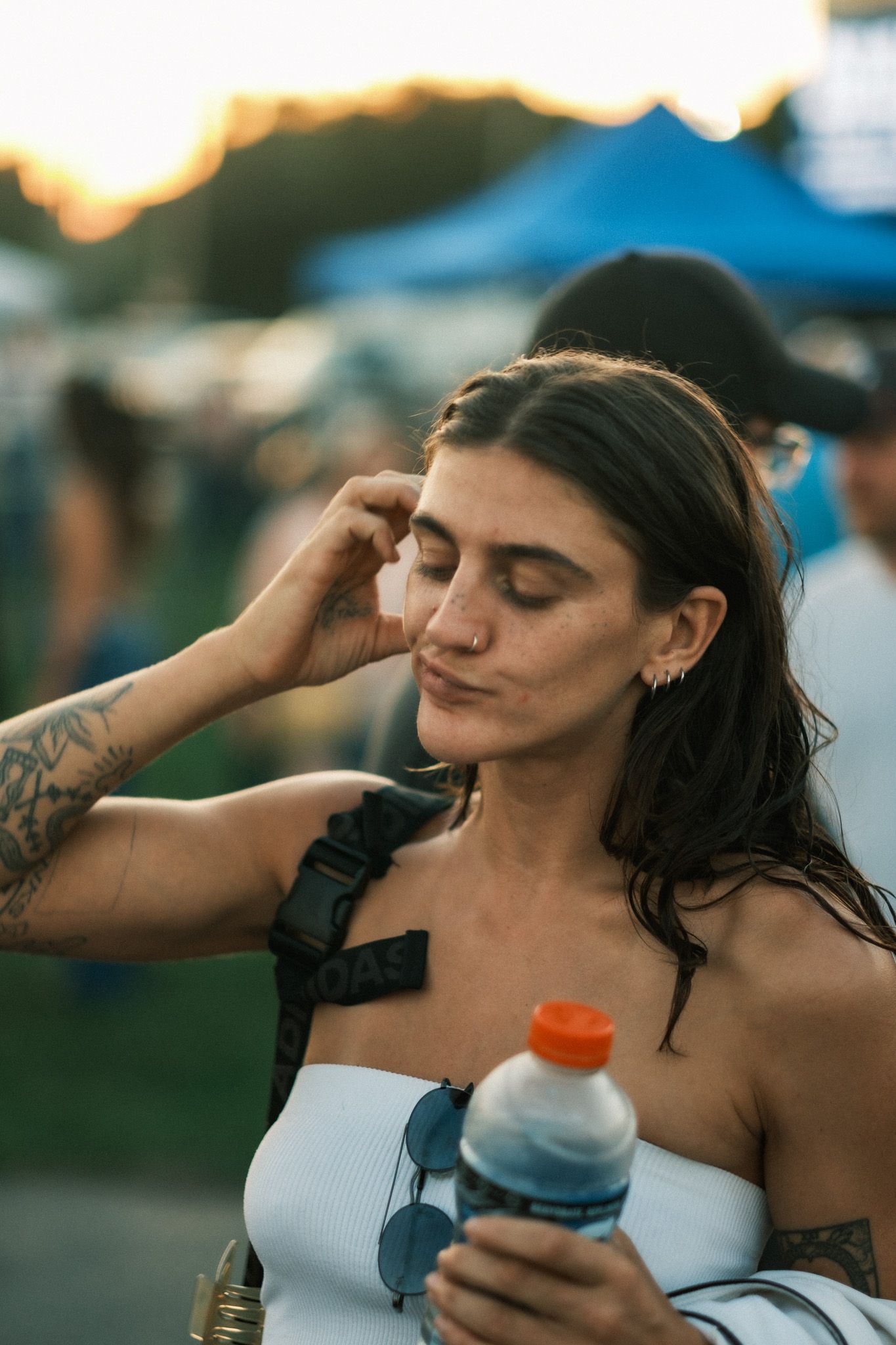 Tattooed woman with dark hair holding a water bottle at an outdoor festival during golden hour sunset.