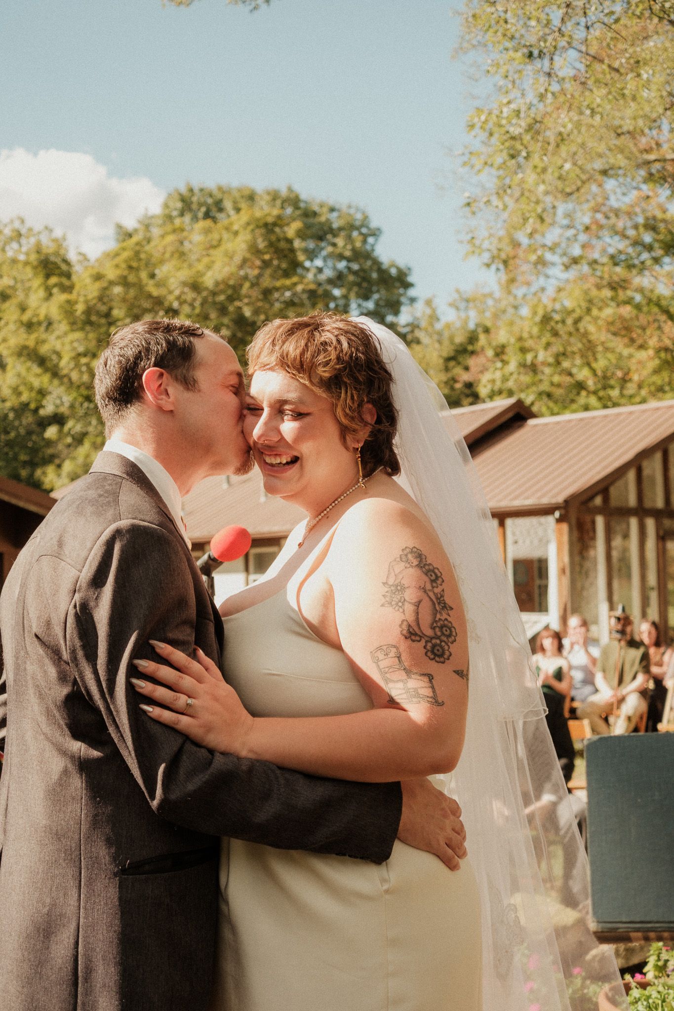 Groom kisses smiling brides cheek during outdoor wedding ceremony with guests in background.