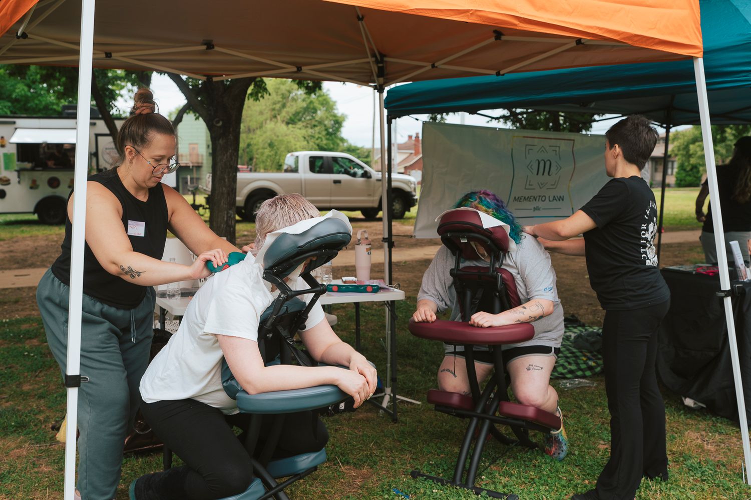Two massage therapists provide chair massages to clients under a tent at an outdoor event.