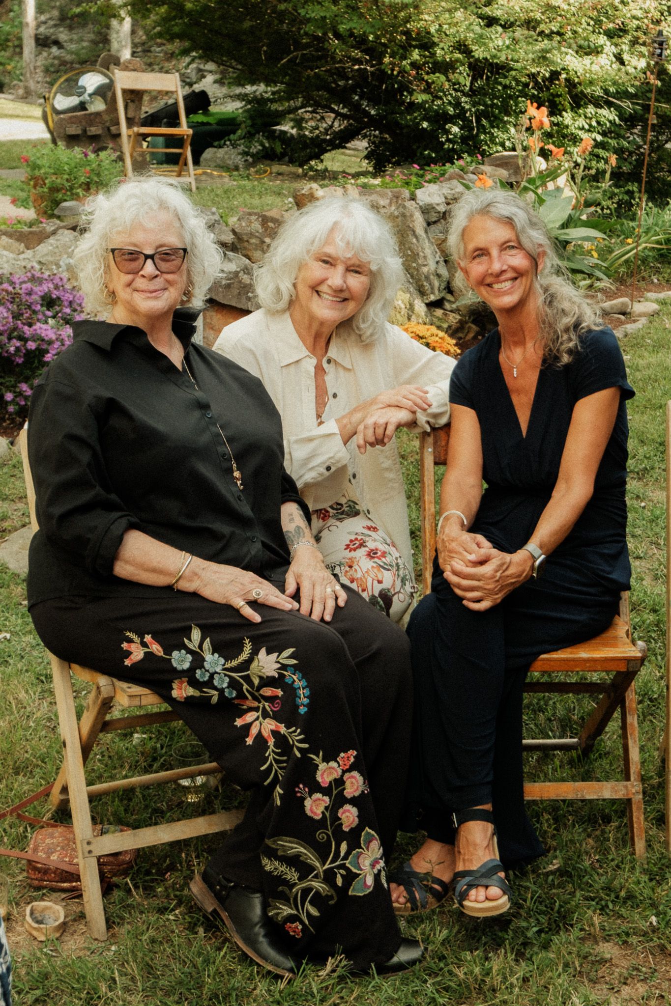 Three elegant older women with white hair sitting together outdoors in a garden setting.