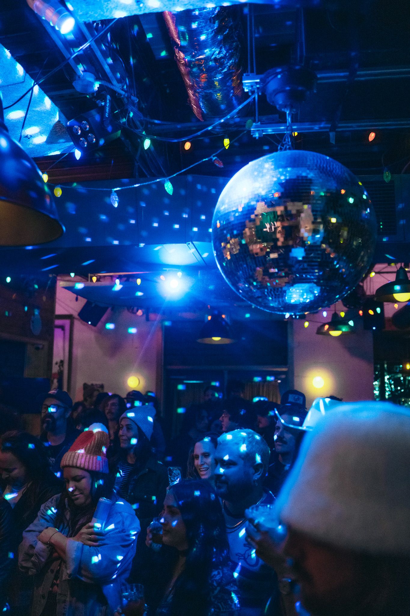 Crowded dance floor glows under a large disco ball with colorful string lights in a dark venue.