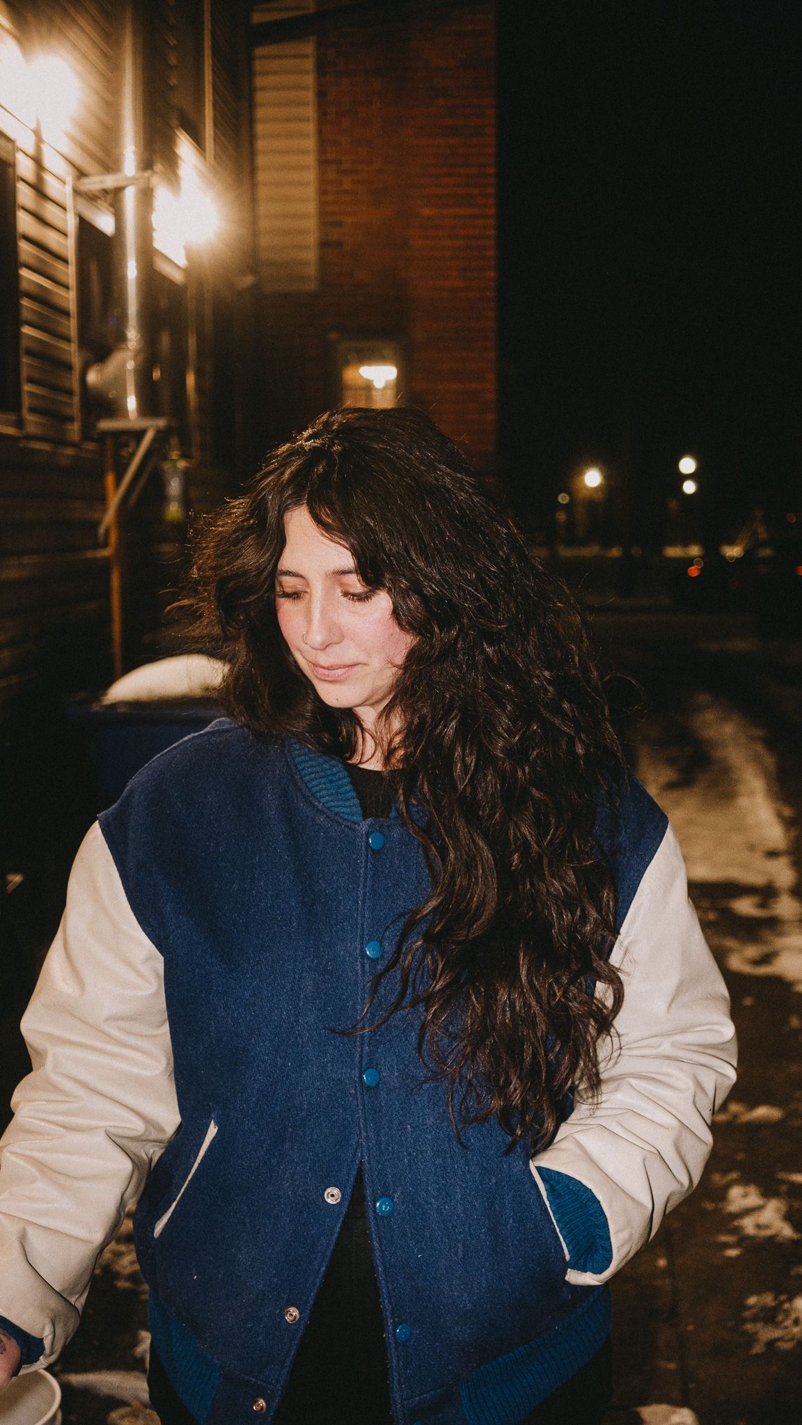Young woman with long curly hair wearing a blue varsity jacket standing outside at night.