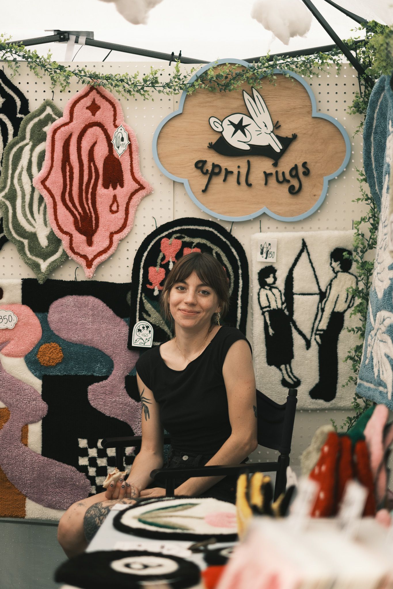 Tattooed woman sitting in front of colorful handmade rugs at an April Rugs craft market booth display.
