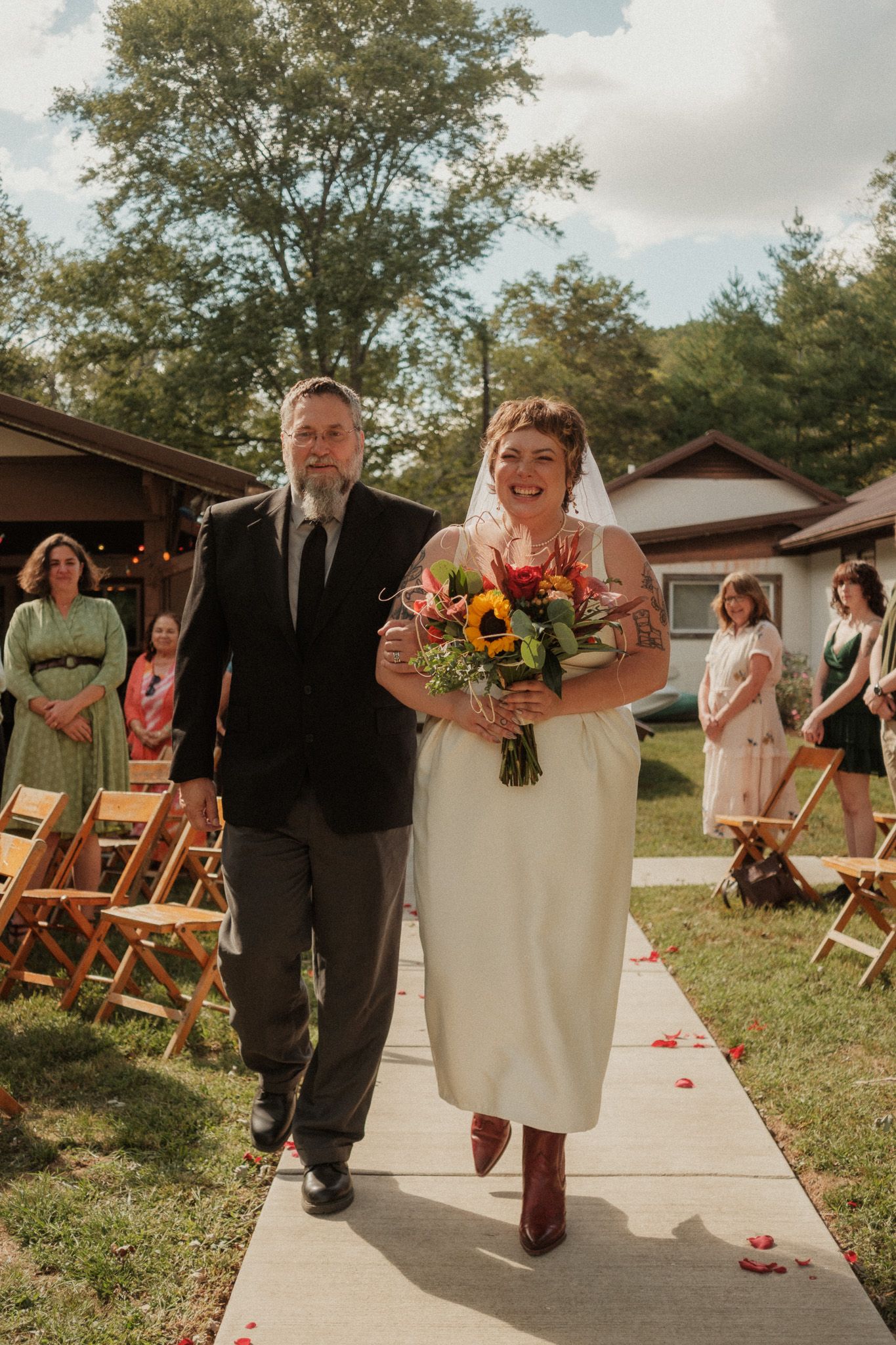Bride walks down outdoor aisle in white midi dress holding sunflower bouquet, escorted by bearded man in black suit.