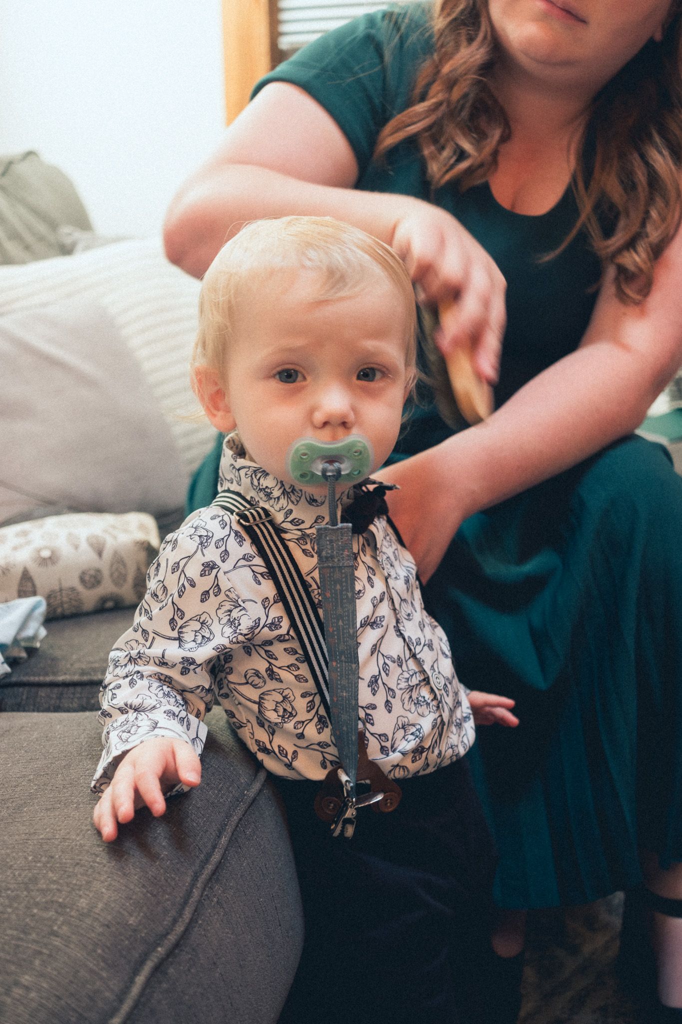 Adorable baby boy with a pacifier wearing a floral shirt with suspenders on a sofa.