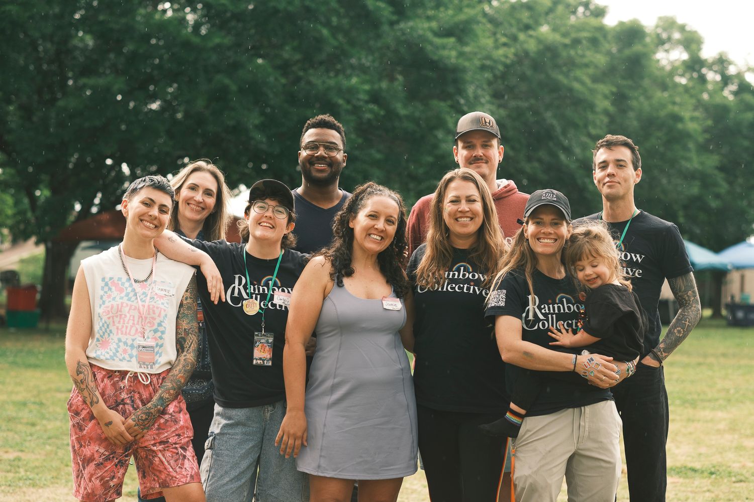 A diverse group of nine people pose together outdoors wearing Rainbow Collective event staff shirts.