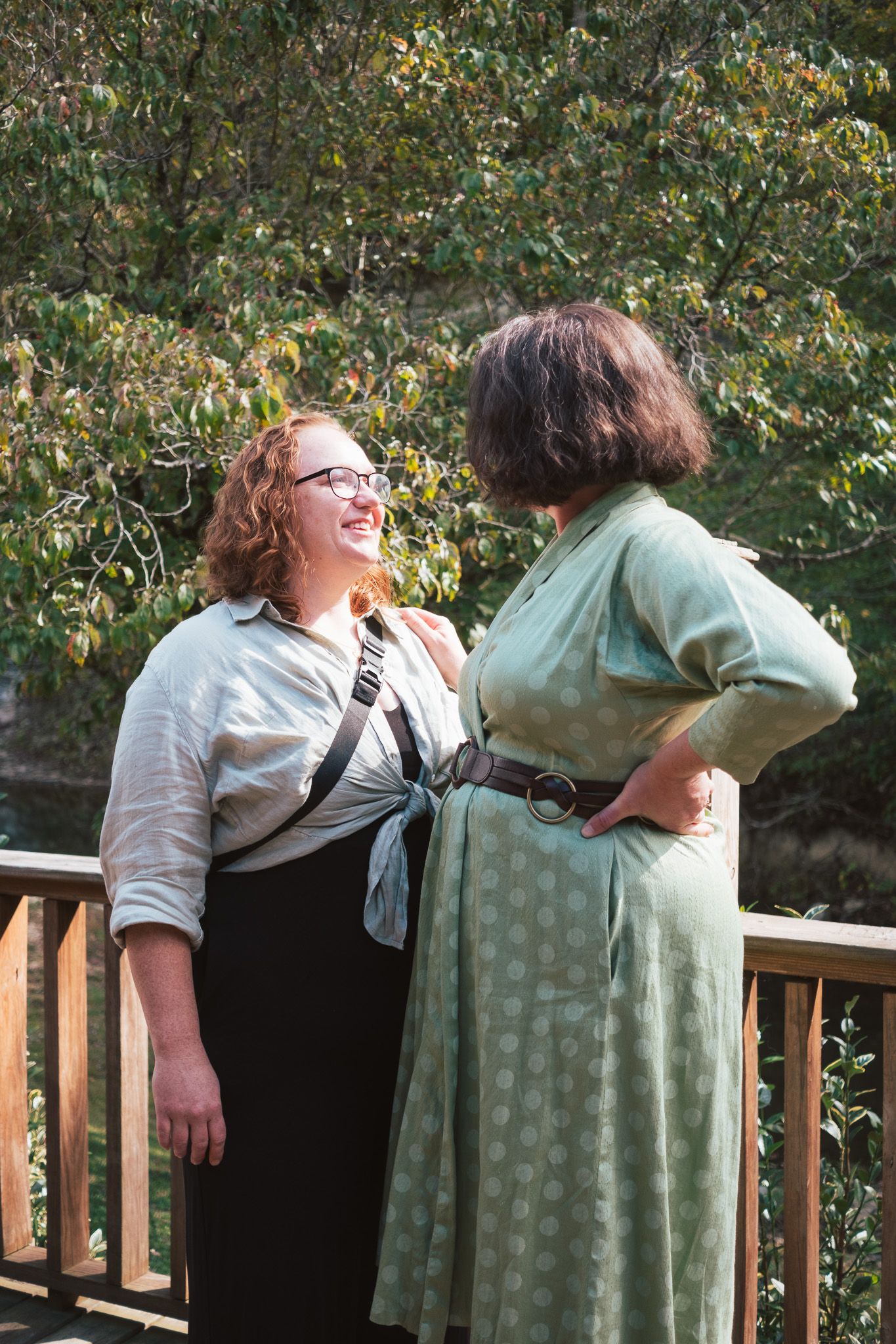 Two women laugh together on wooden deck surrounded by green trees at outdoor wedding.