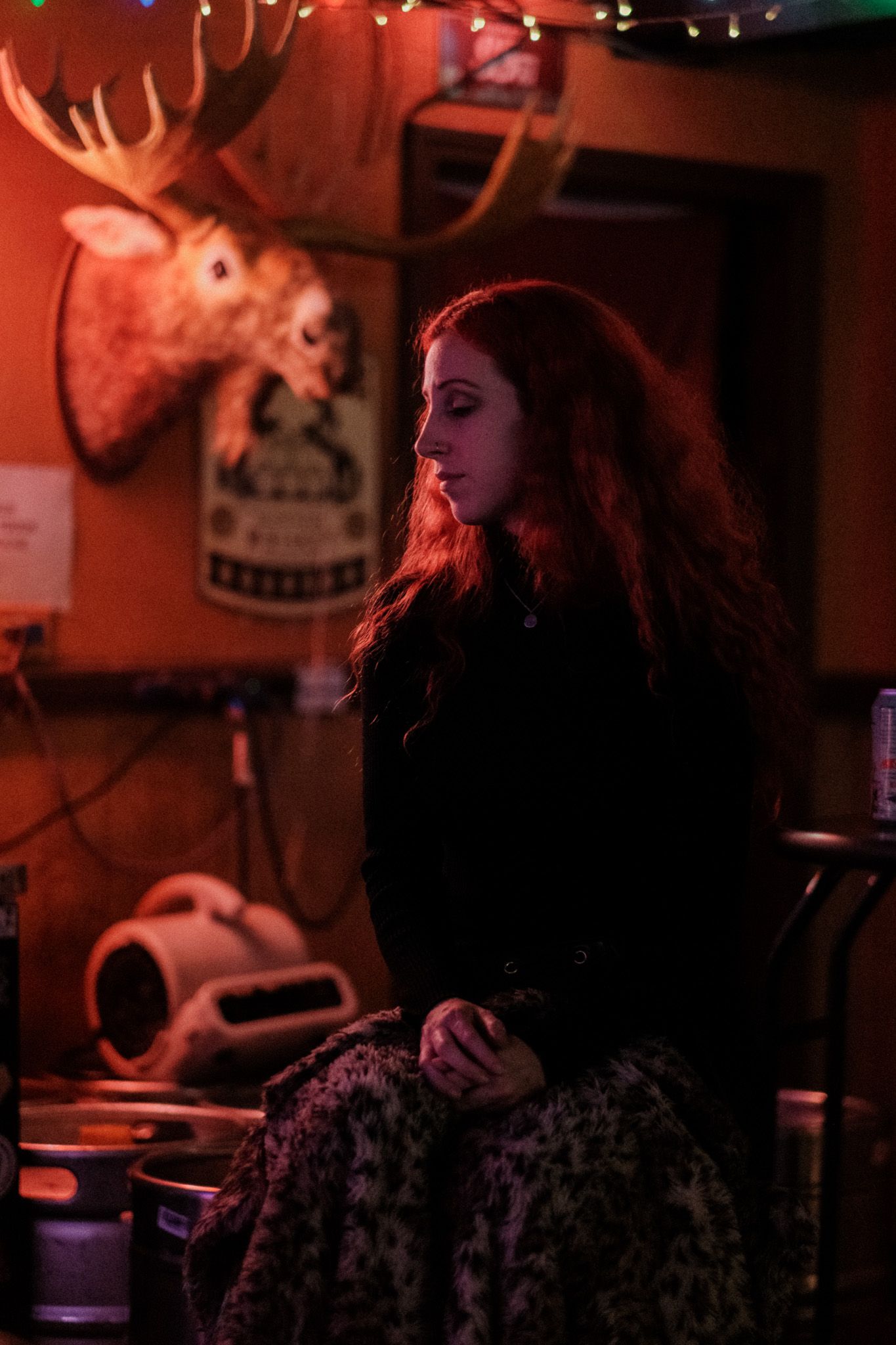Young woman with long red hair sitting in a dimly lit bar with moose head mounted on wall.