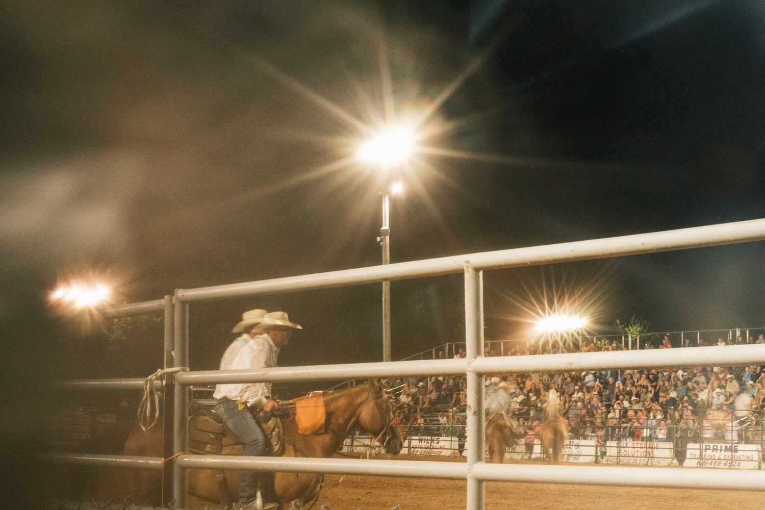 Cowboy riding a horse in a rodeo arena illuminated by bright stadium lights at night.