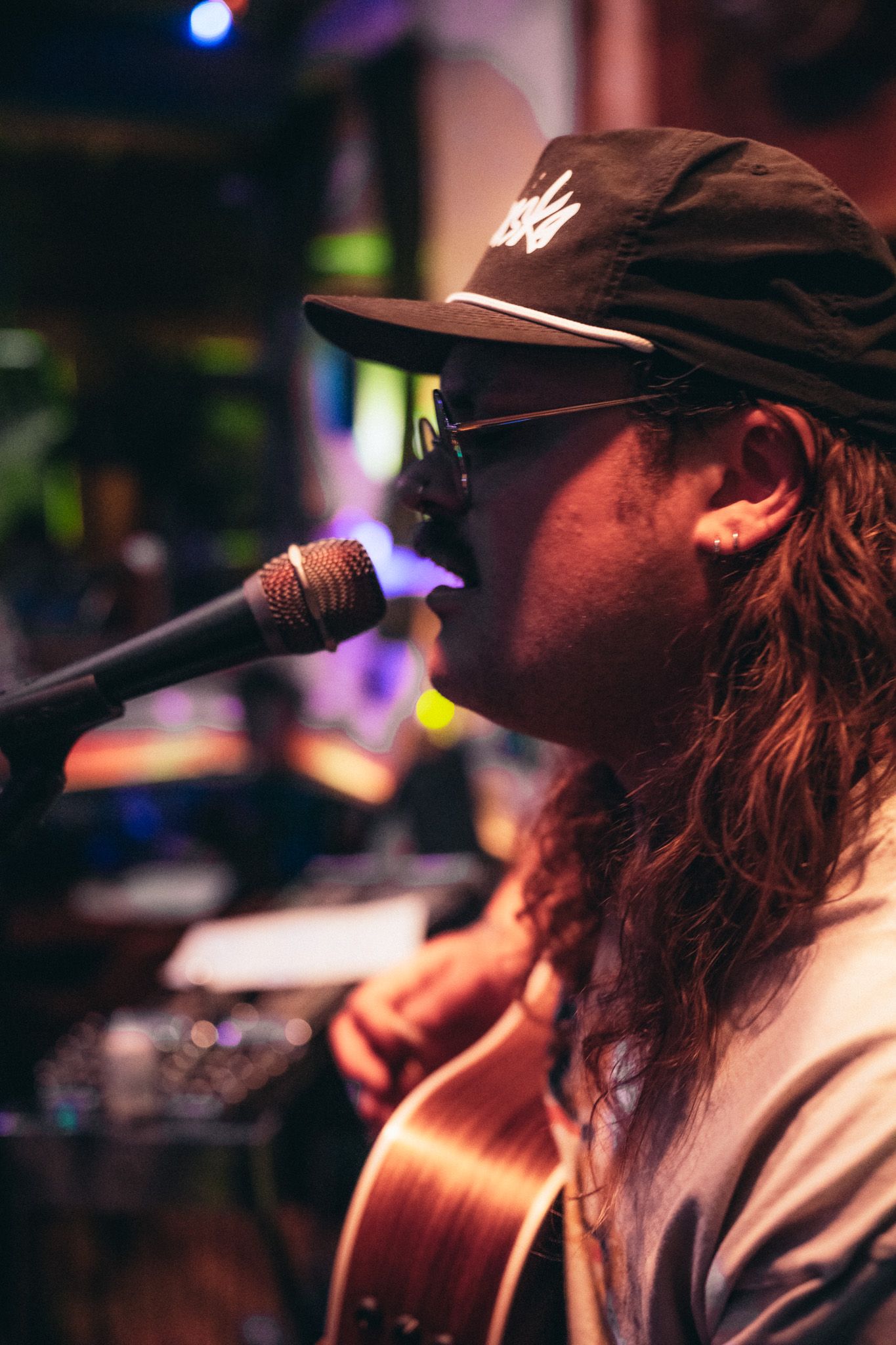 Long-haired guitarist wearing a black cap and sunglasses singing into a microphone during a live performance.