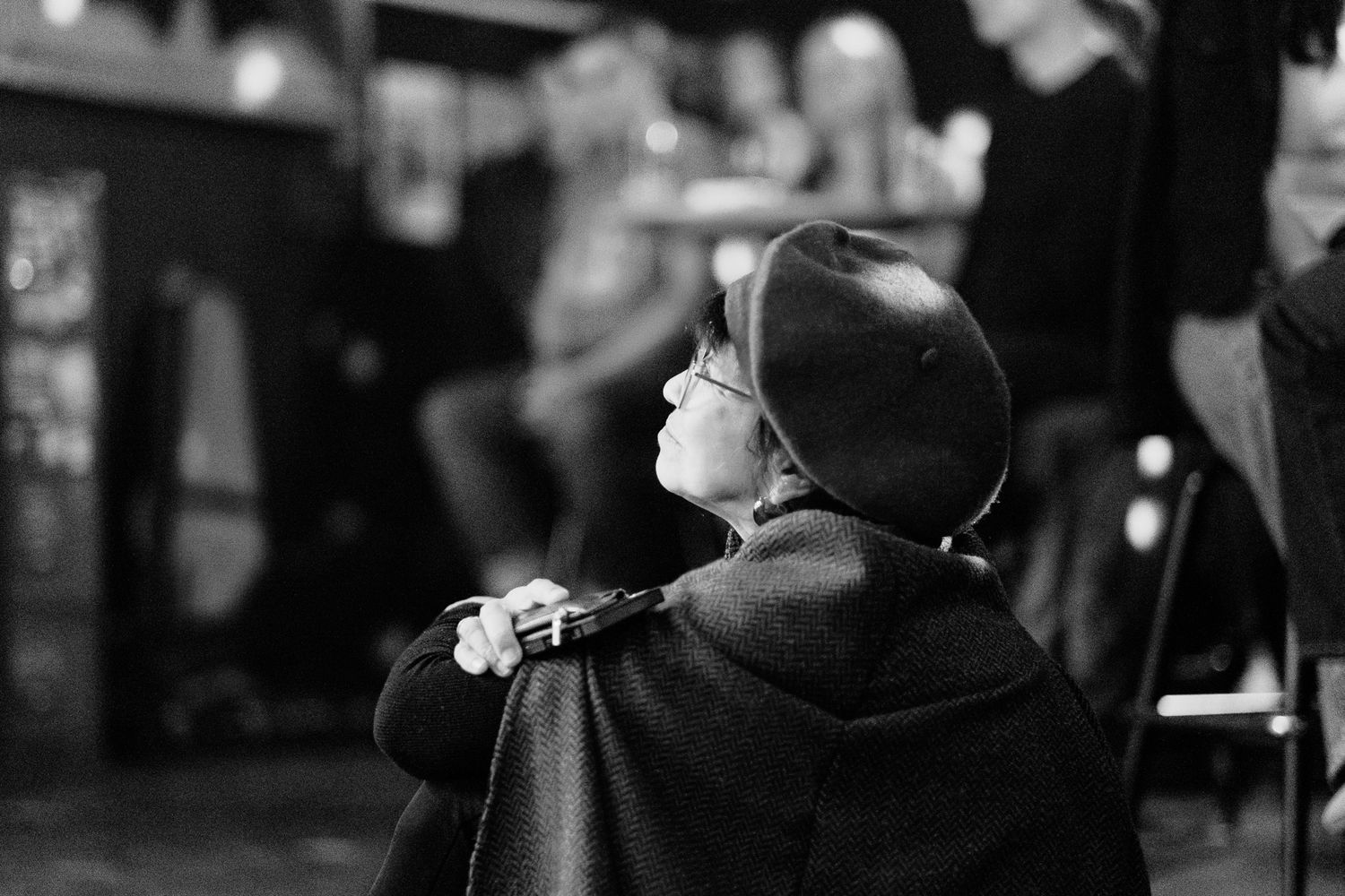 Black and white photo of an elderly person wearing a beret and cape sitting in a busy cafe.