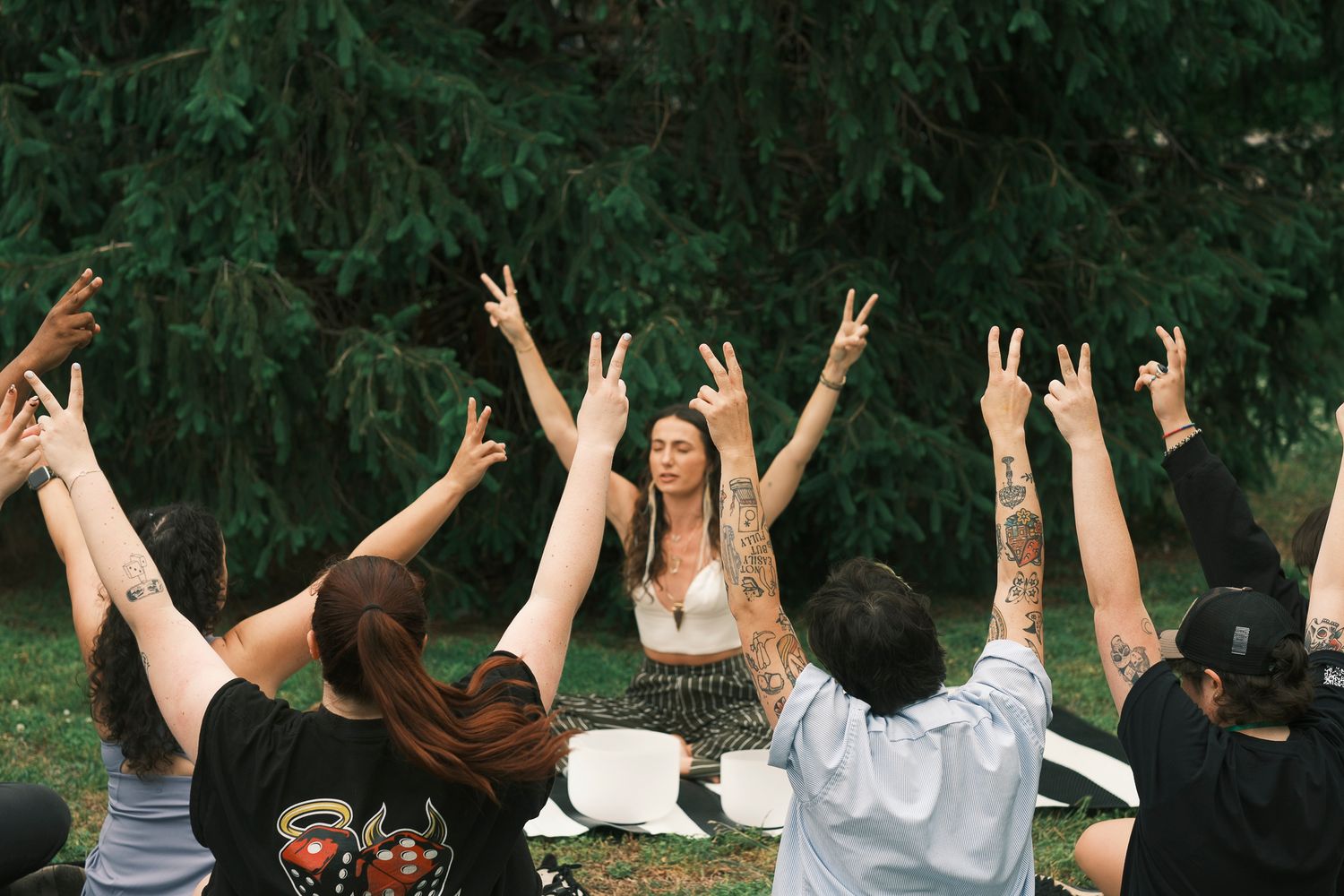 A group of people raise their hands in peace signs during an outdoor yoga session under pine trees.