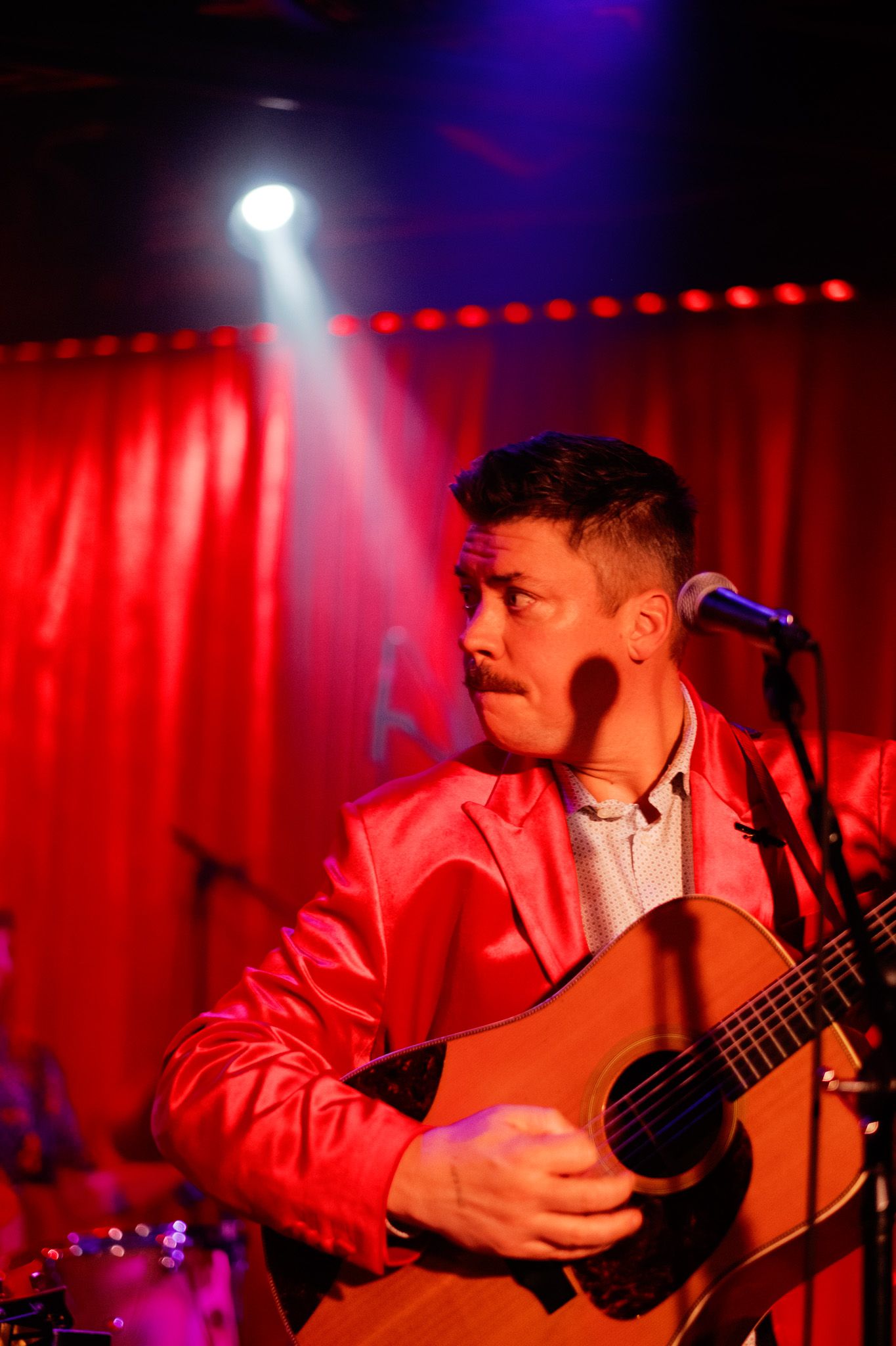 Male guitarist performing on stage in a red jacket under dramatic red and blue stage lighting.