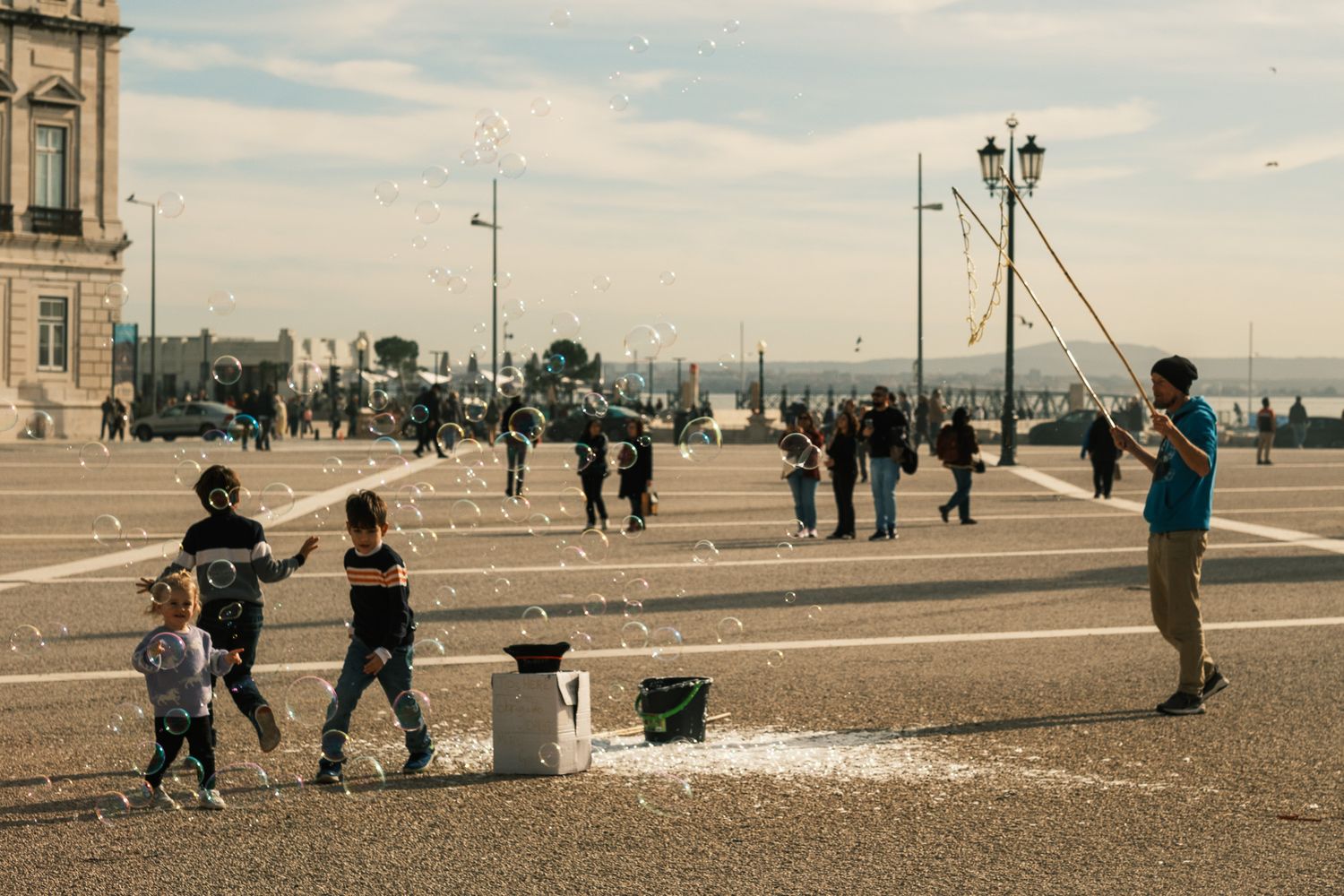 Street performer creating giant soap bubbles for children in a large outdoor plaza.