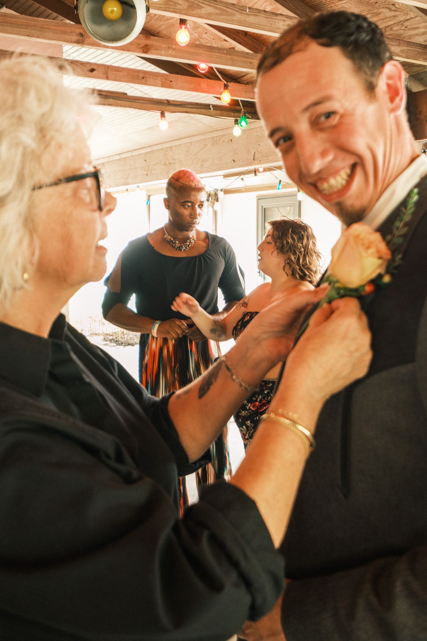 Woman pins boutonniere on smiling groom's lapel while guests socialize at rustic indoor wedding venue.