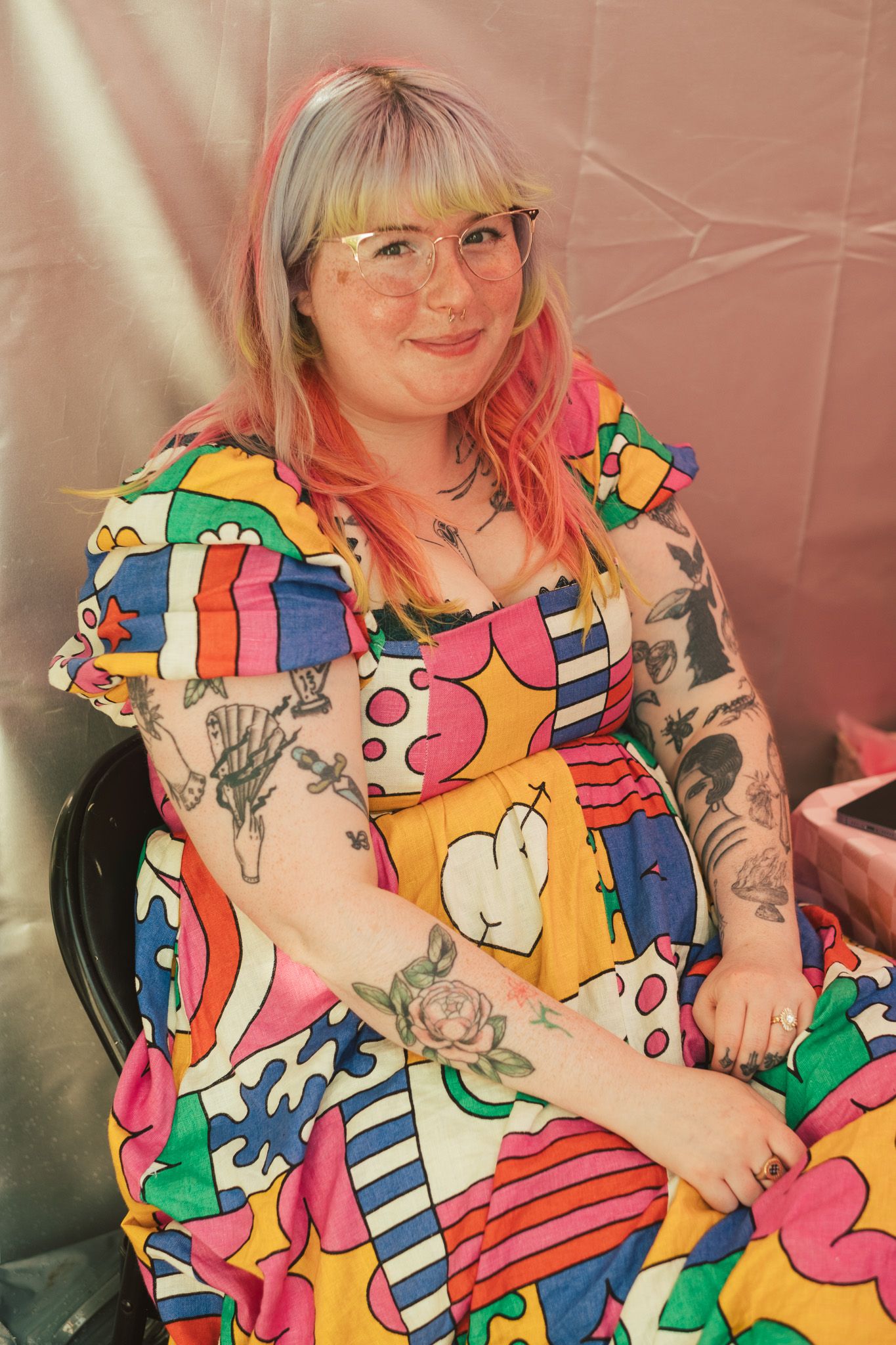 Plus-size woman with pink hair and tattoos wearing a vibrant multicolored dress, sitting and smiling warmly.