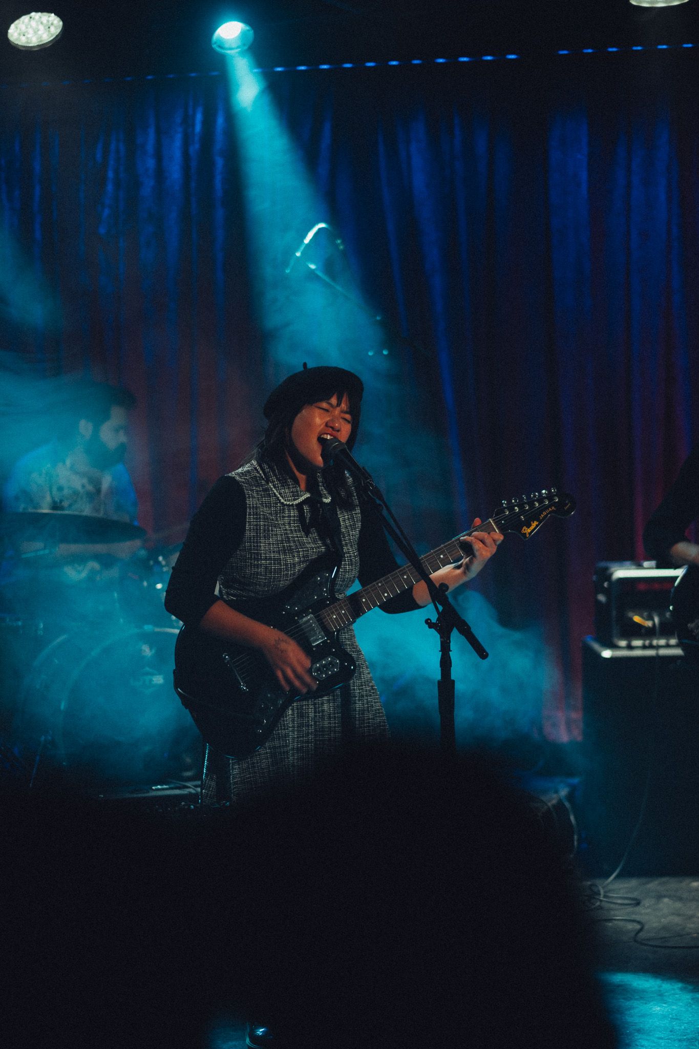 Female guitarist in beret smiling while performing on stage under blue spotlights with smoke effects.