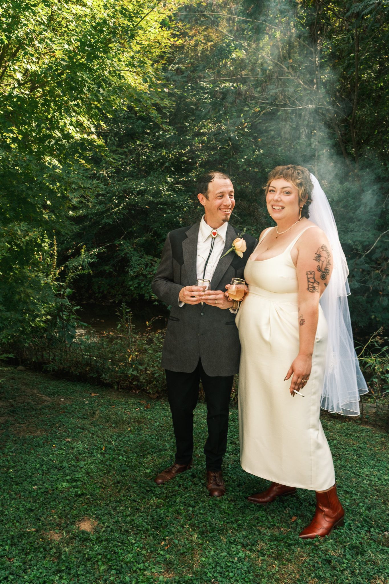 Bride and groom smiling outdoors in lush green garden, bride wearing white dress with veil and boots.
