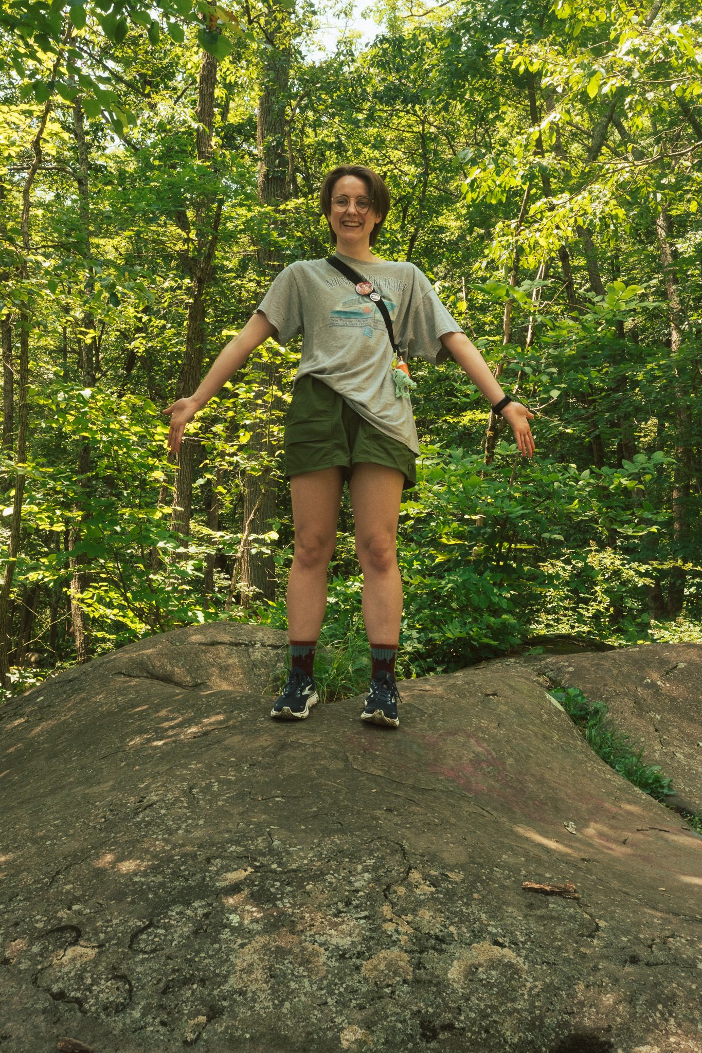 Smiling woman standing on a large rock in a lush green forest wearing green shorts and a gray t-shirt.
