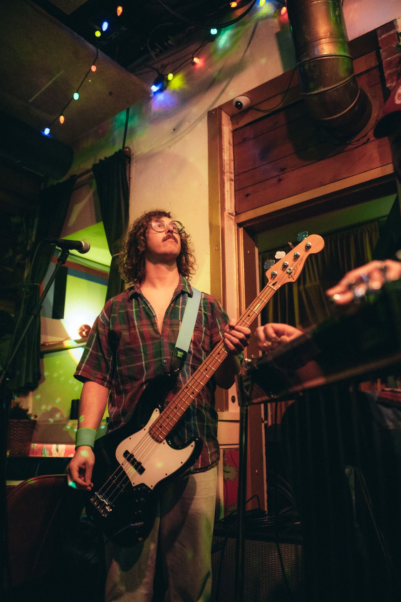Curly-haired musician plays bass guitar on a dimly lit stage with colorful string lights overhead.