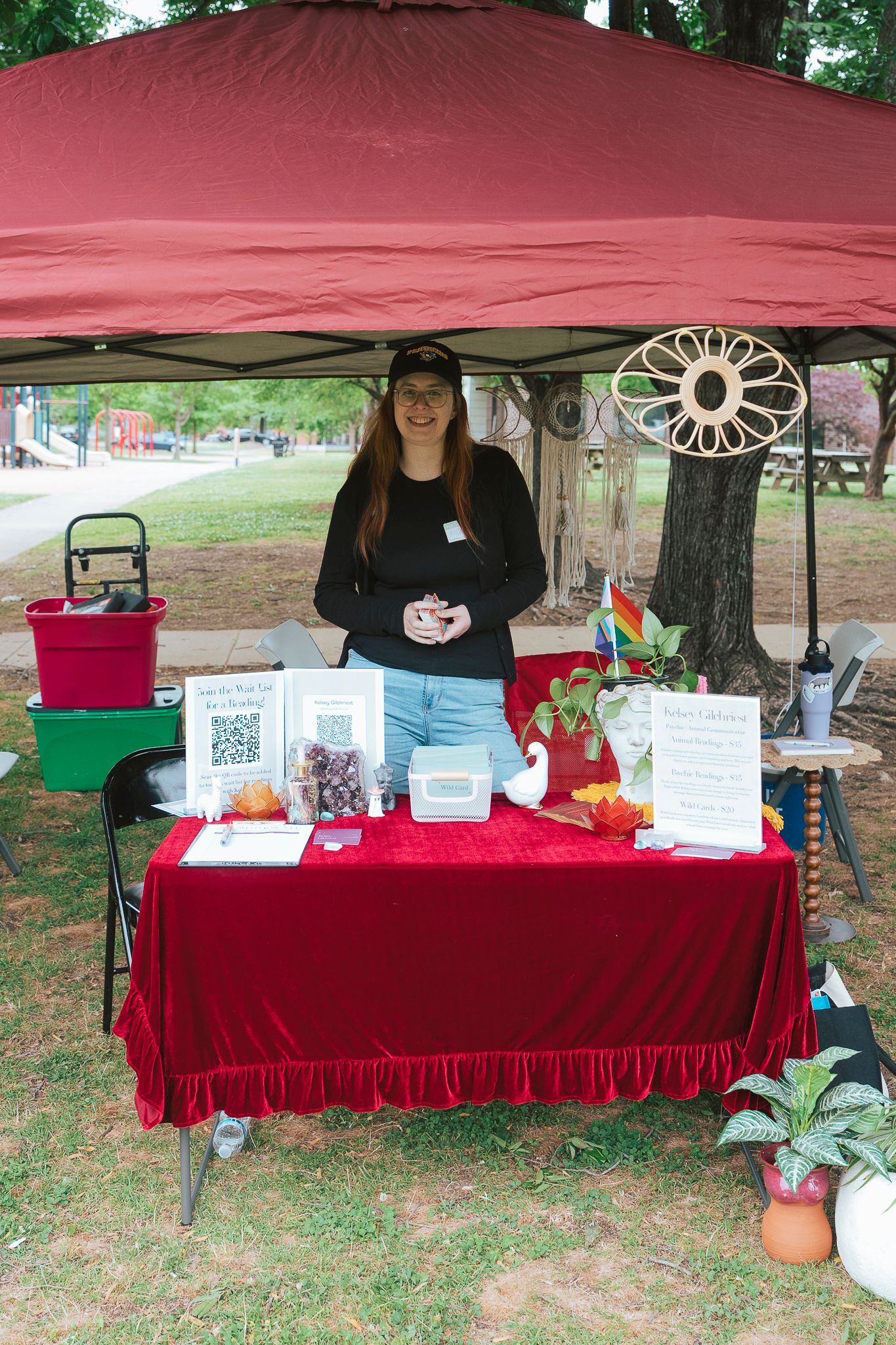 Young woman standing at red velvet table display booth with crystals and plants at outdoor market.